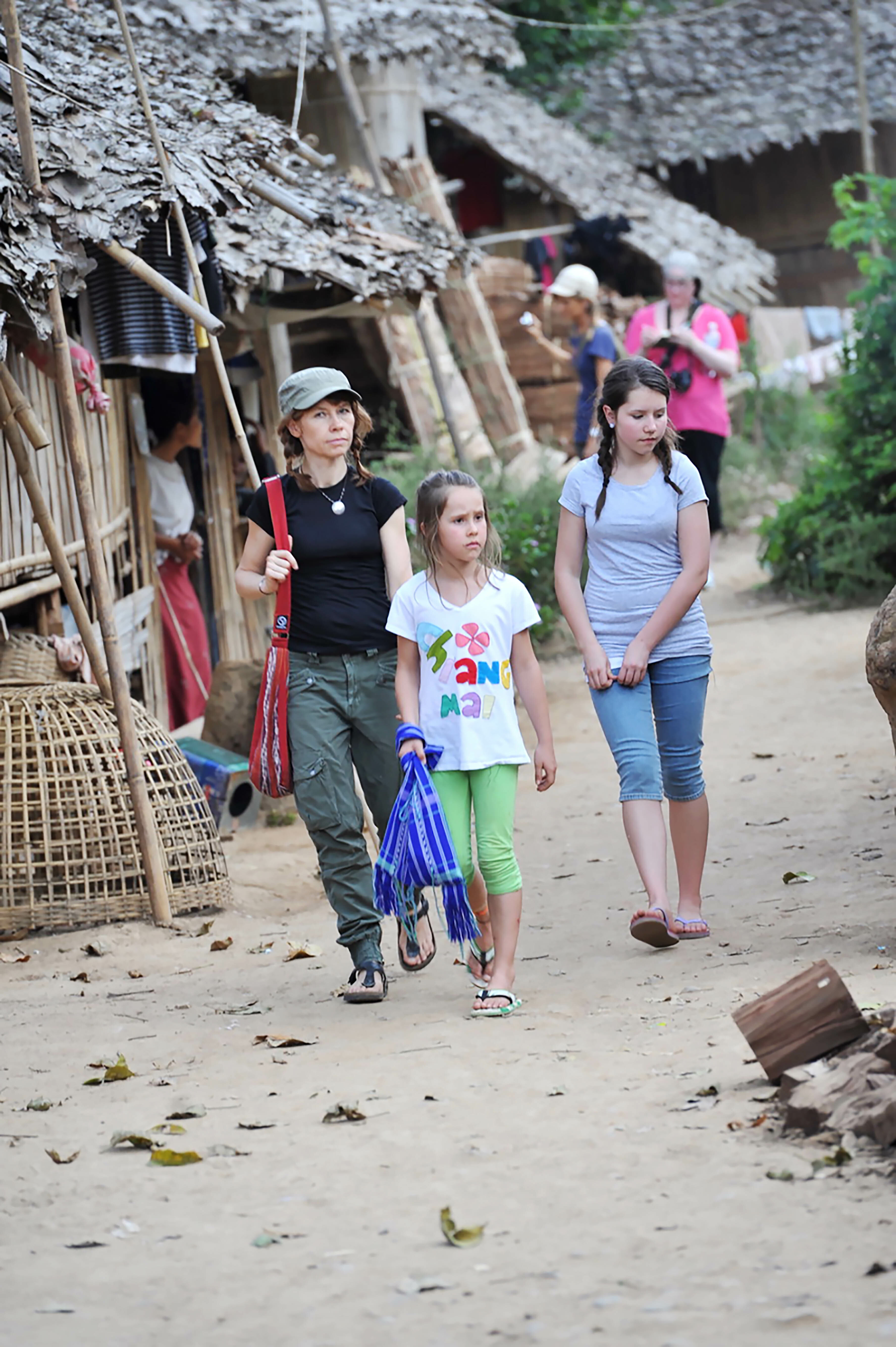 Foto: en kvinne i grønn bukse, caps og svart t-skjorte går mellom enkle hus med tak som ser ut som de er laget av en type bark. Hun har med seg to henter i t-skjorte og knelange tights.