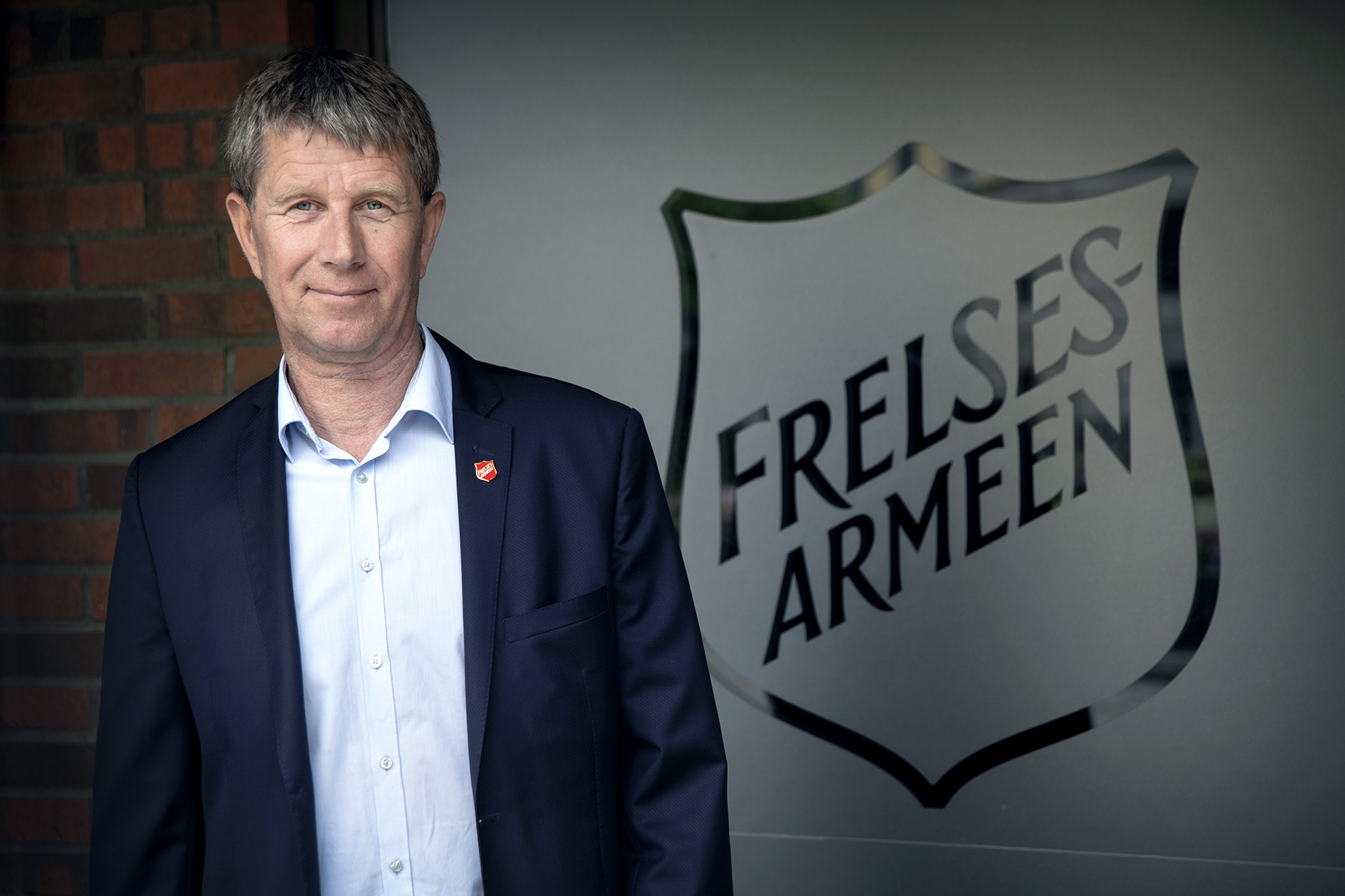 Foto: Mann i 60-årene i lyseblå skjorte og blå dressjakke står inntil en frostet glassrute med Frelsesarmeens skjold. han har også på seg en rød pin med logoen på jakkekragen.