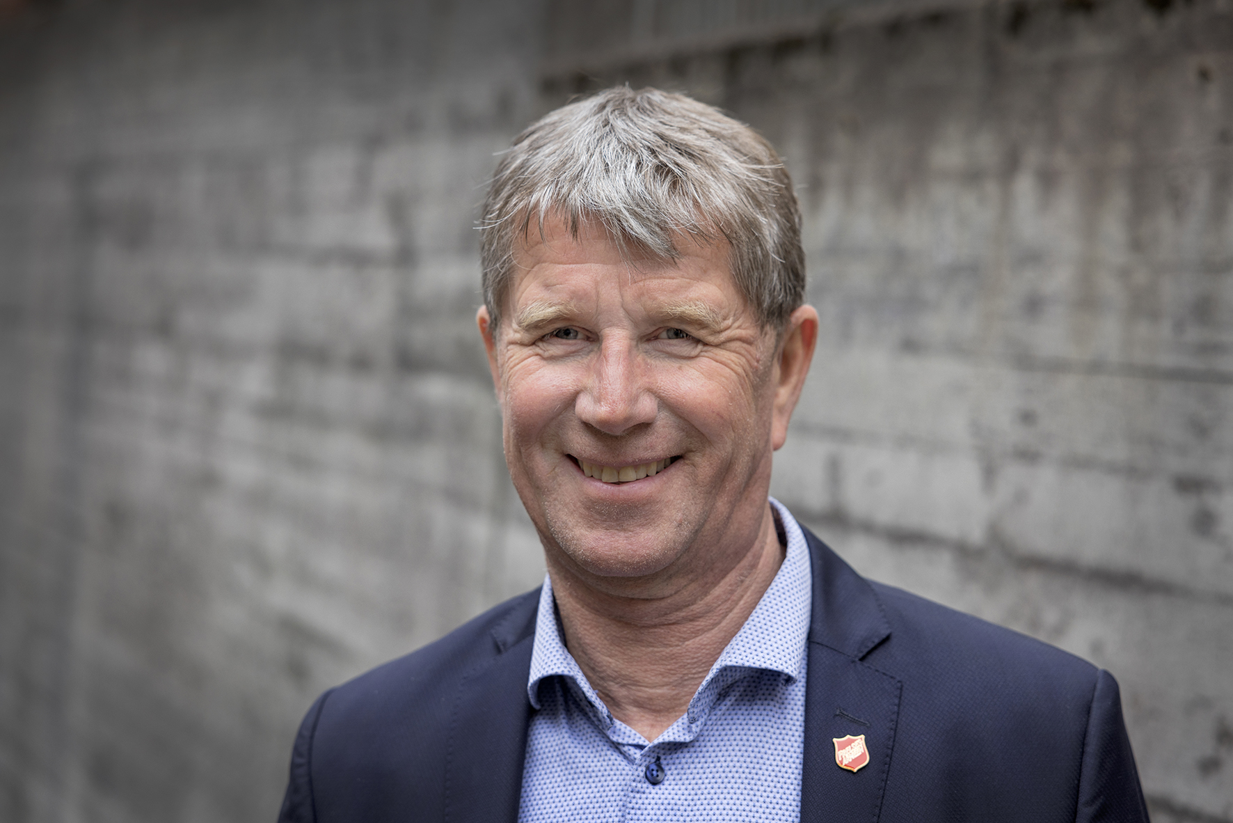 Foto: Mann i 60-årene står ute, mot en betongvegg. Han har på seg blå skjorte og mørkeblå dressjakke. Han smiler og har grått hår.