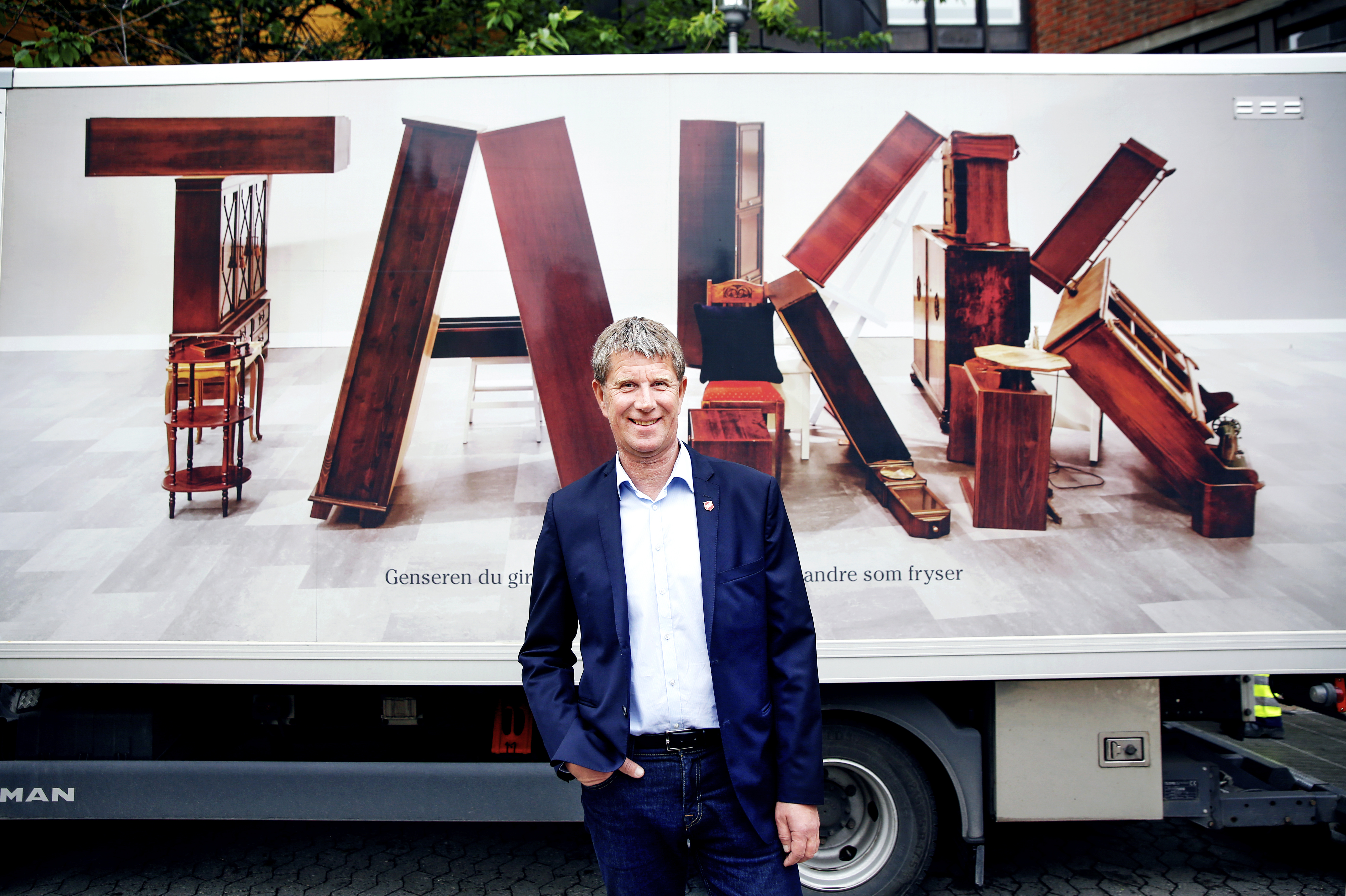 Mann i skjorte og dressjakke står foran en hvit lastebil som er foliert med et bilde av ordet TAKK som er "skrevet" ved at gamle møbler er stablet sammen til fire bokstaver.