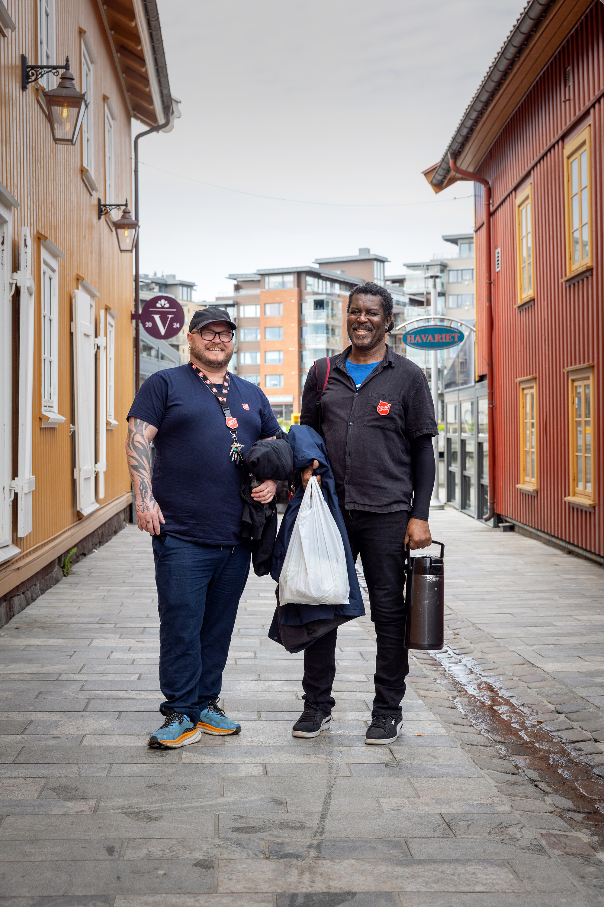To menn, en hvit og en svart, står i en gågate. Begge har på seg blå klær med logo fra Frelsesarmeen.