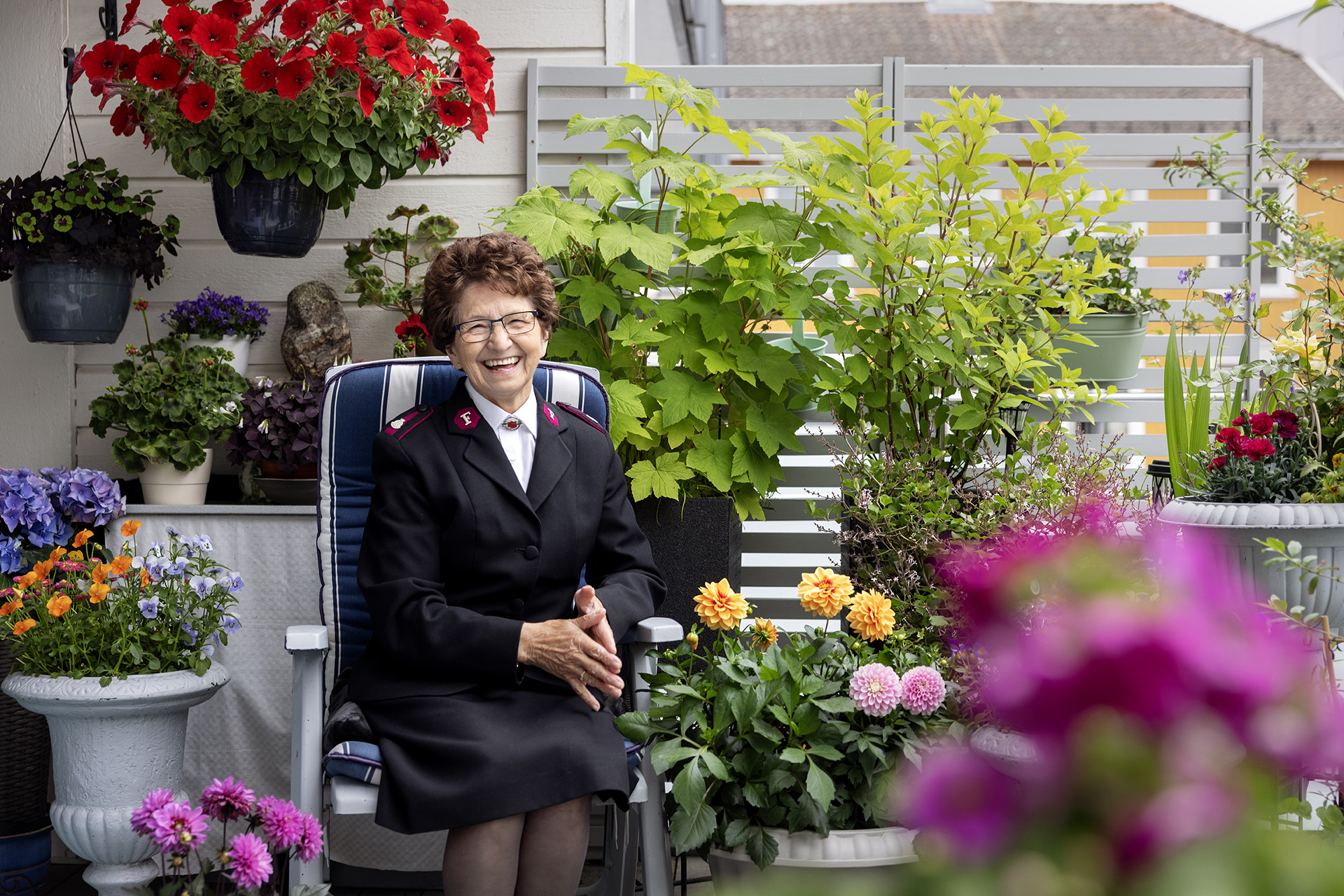 Foto: En kvinne i Frelsesarme-uniform sitter på en balkong og smiler mot kamera. Rundt henne sees planter og blomster i mange farger.