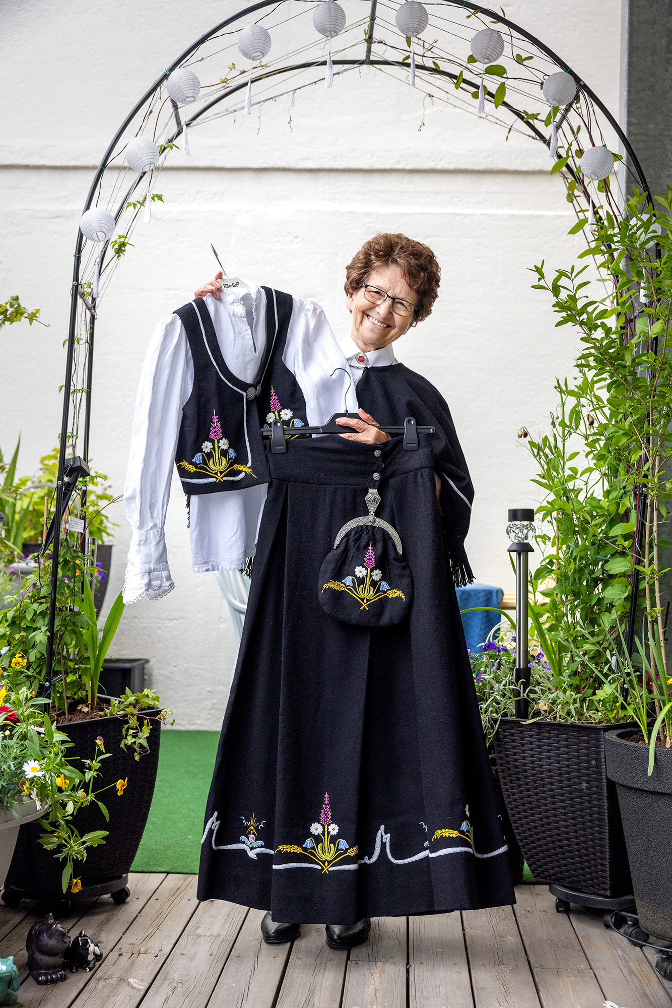Foto: En eldre kvinne står under en hagebue i metall, pyntet med små, runde lys. På begge sider er det planter. Kvinnen holder en svart bunad og hvit bunnadsskjorte foran seg.