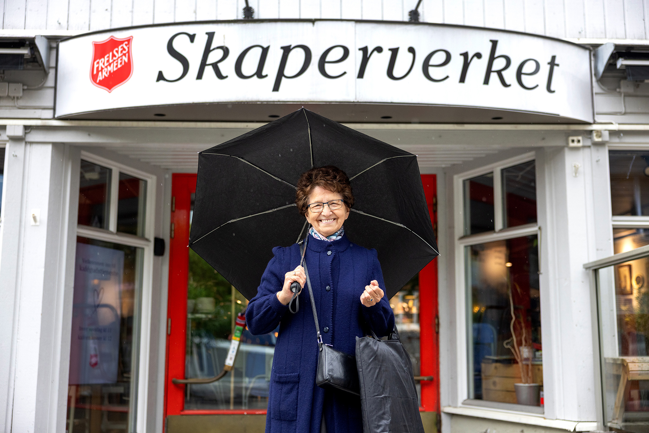 En eldre kvinne med brunt, kort hår og briller står under en svart paraply og smiler. Bak henne er et hvitt trebygg med et stort skilt der det står Skaperverket.
