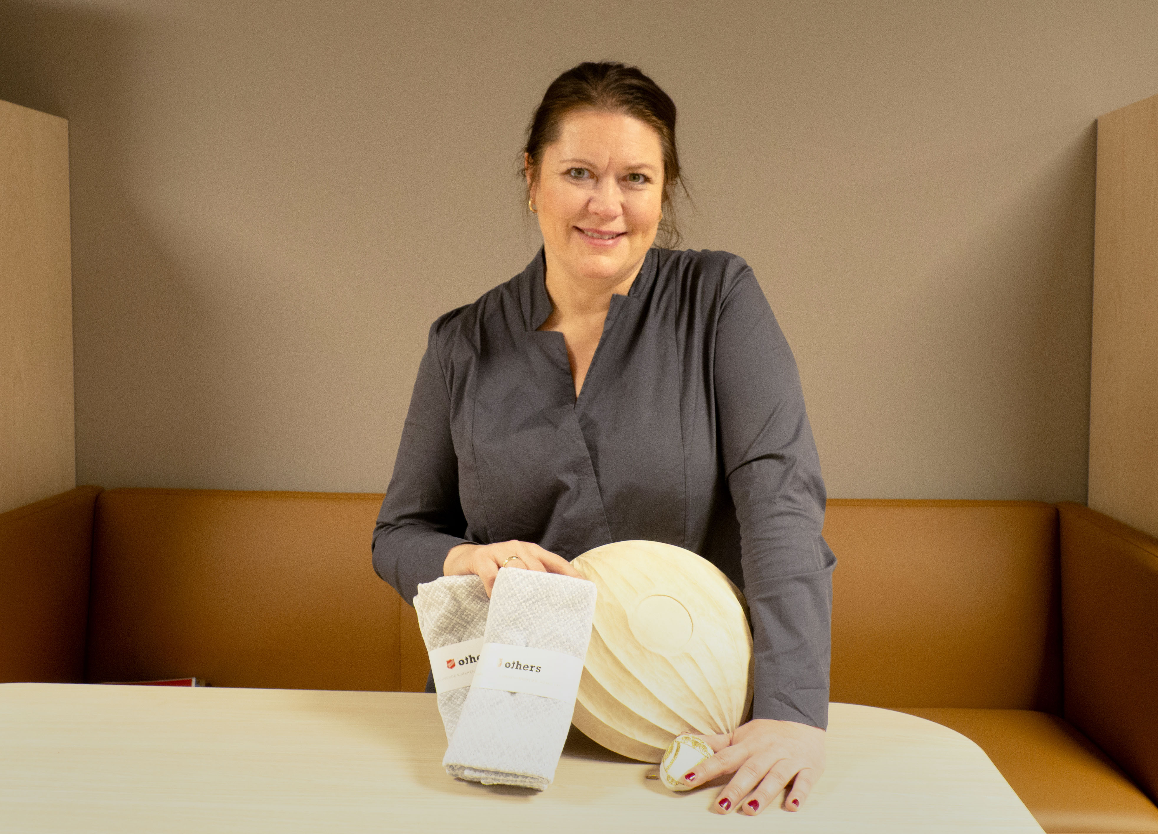 Foto: en kvinne med brunt hår i hestehale og blågrå skjorte står ved et bord, på bordet foran seg har hun en trebolle og to sammenbrettede kjøkkenhåndklær.