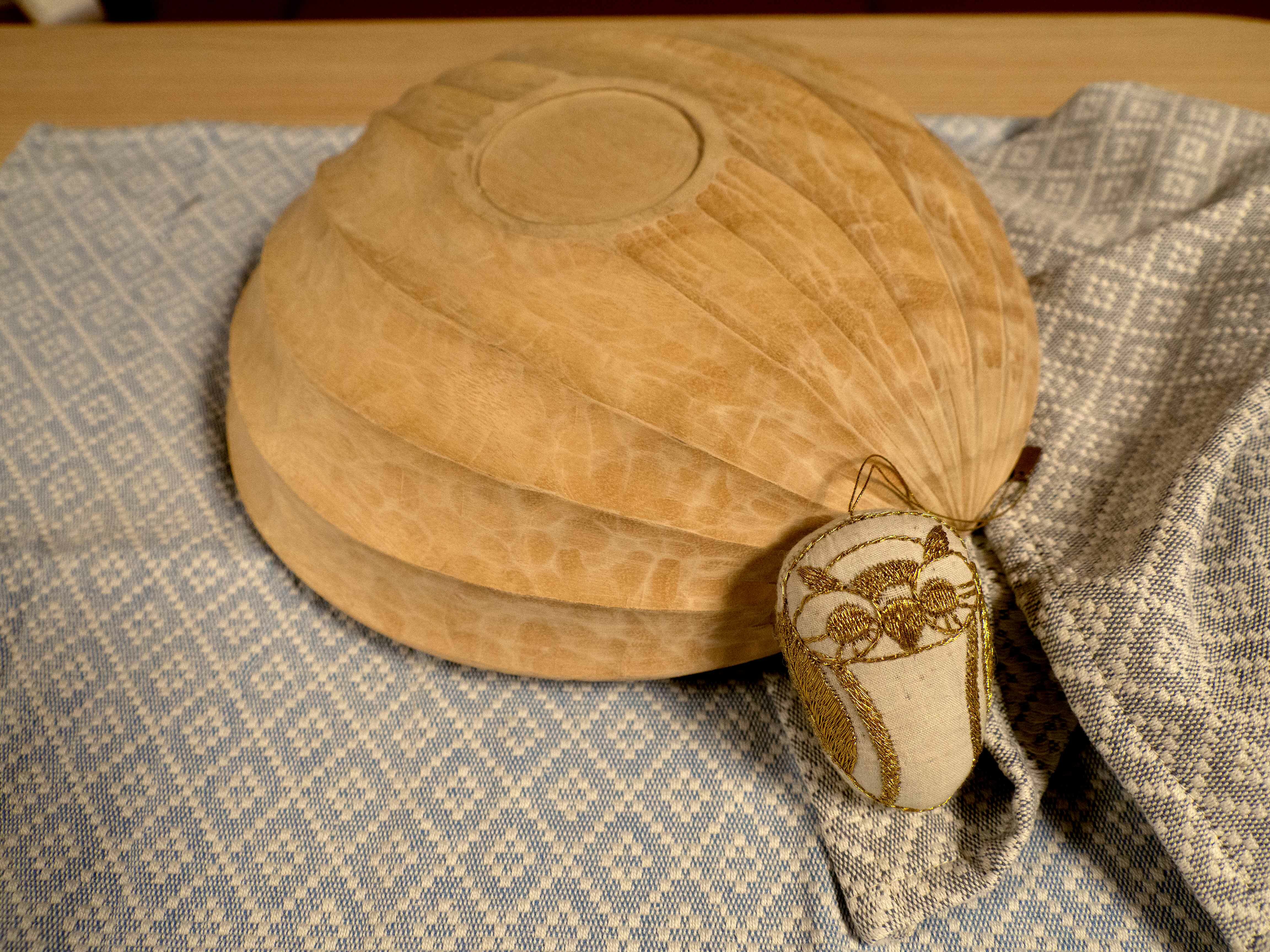 Foto: En trebolle ligger opp ned på et vevd håndklestoff med repeterende prismeformet mønster i grått og beige. Bollen har tydelige skårne striper og minner om bunnen på en båt. Foran ligger en liten brodert ugle i beige med gulldetaljer.