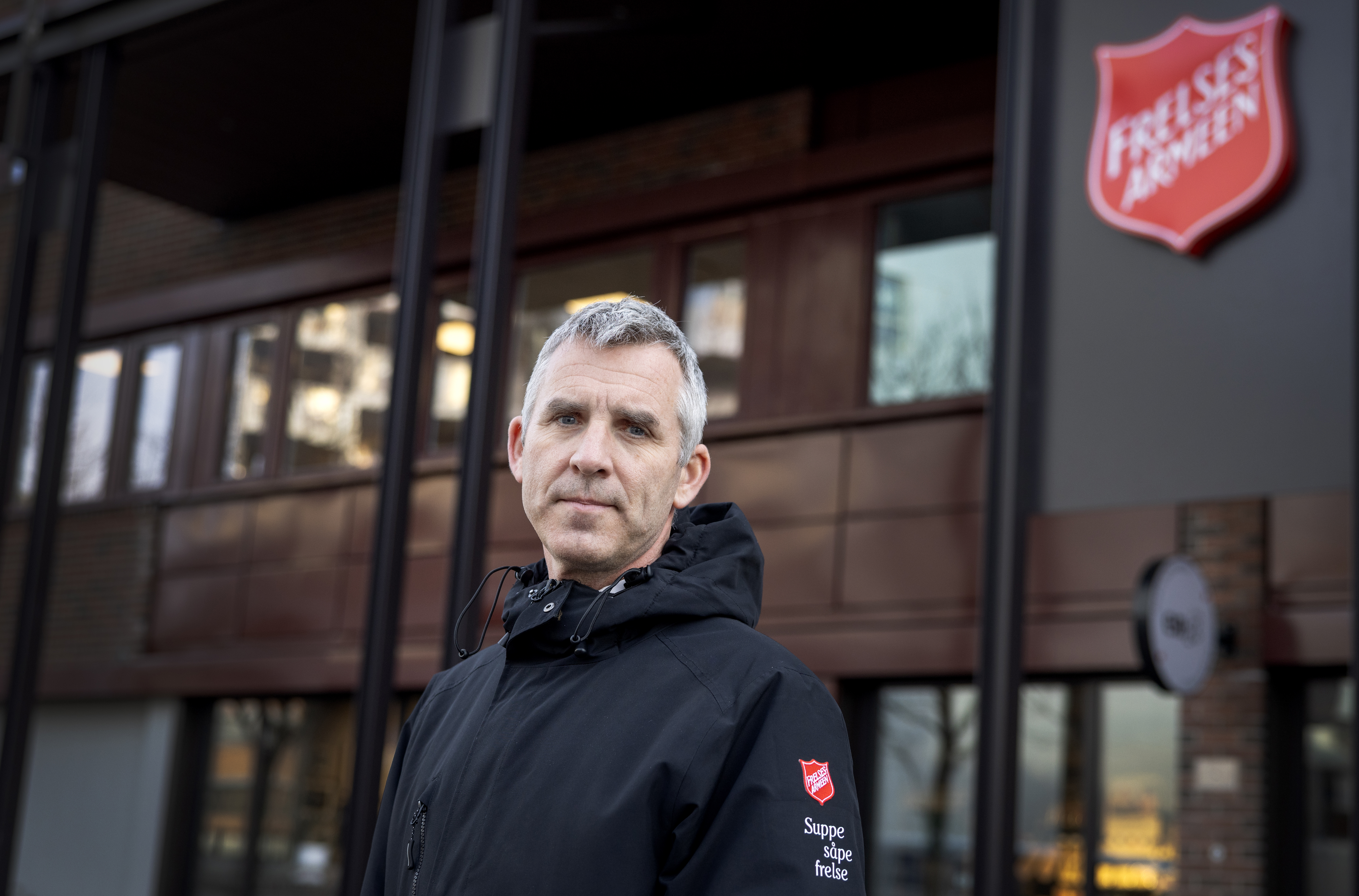 Foto: Mann i 50-årene med svart jakke med rød logo fra Frelsesarmeen. Han står foran et bygg med samme logo.