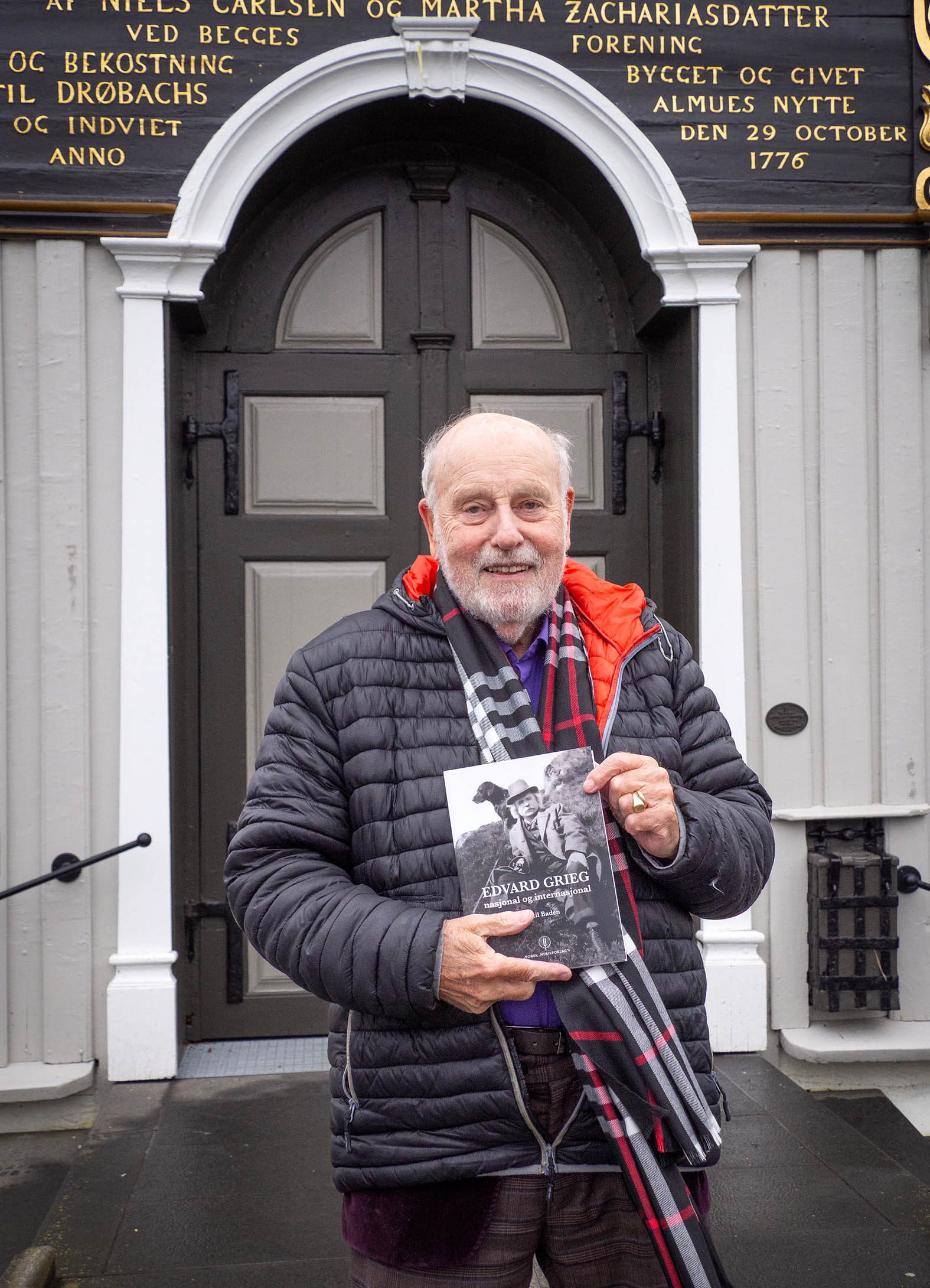 Foto: Mann i 80-årene står utenfor et utsmykket inngangsparti til et gammelt hus. Han holder opp en bok med et svart-hvitt bilde på cover.