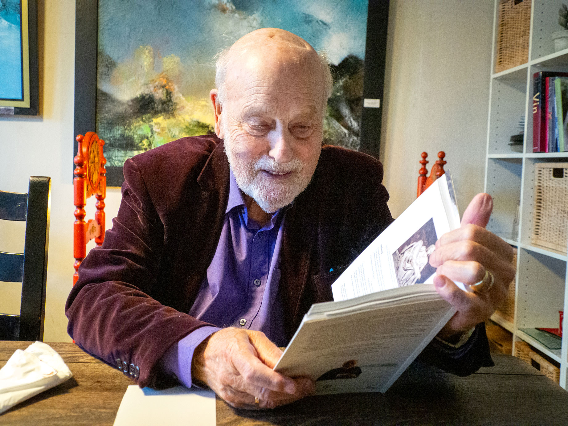 Foto: Manni 80-årene med blank isse og grått skjegg ser ned i en bok som han leser fra. Han sitter ved et trebord og der er bøker i en bokhylle ved siden av og malerier på veggene. Han har på seg lilla skjorte og burgunder dressjakke i velur.