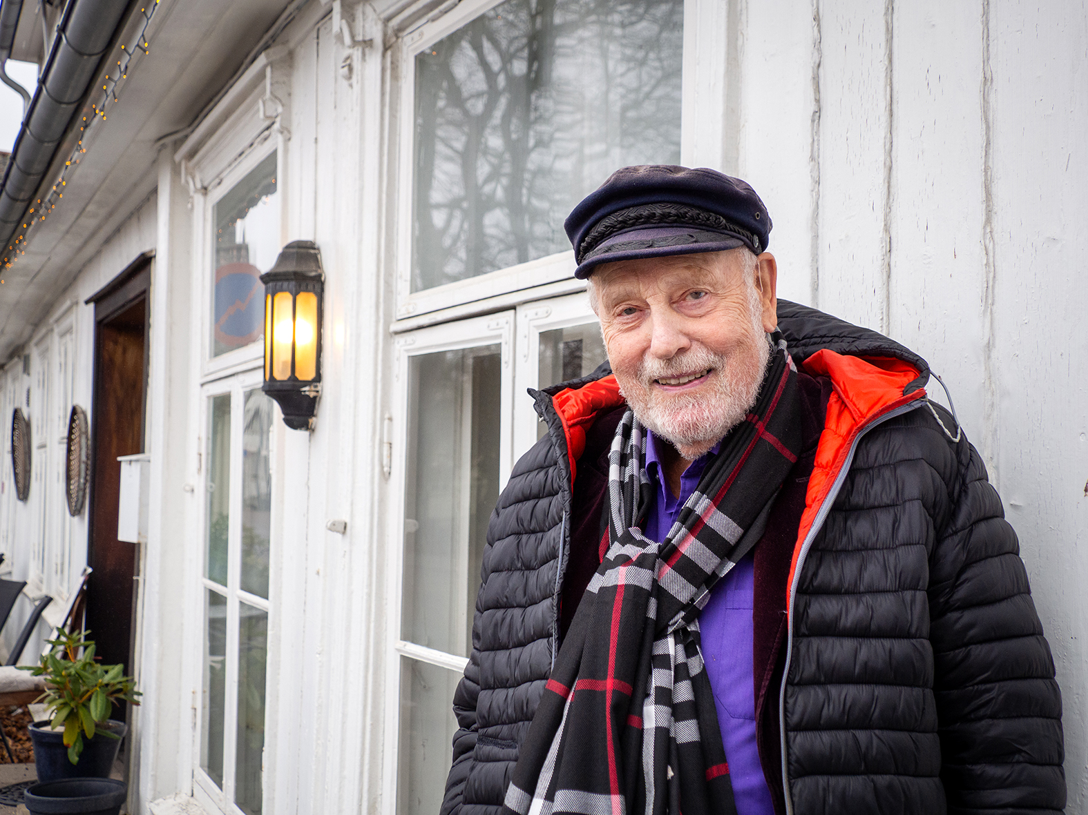 Foto: Mann i 80-årene med skipperlue, skjerf og boblejakke står foran en hvit vegg trevegg med hvite vinduskarmer.