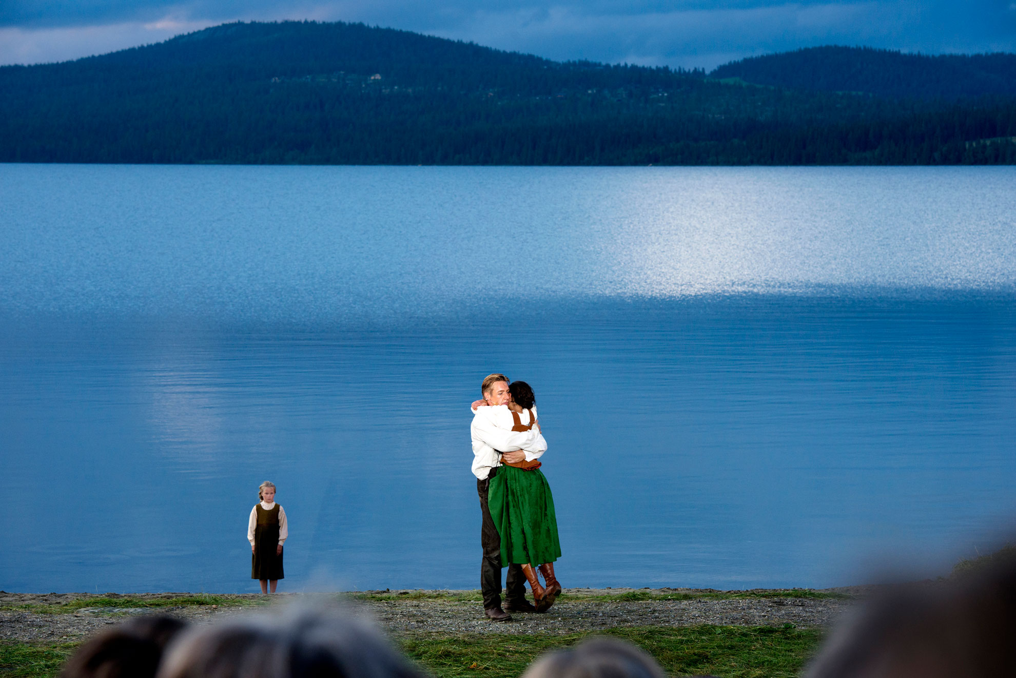 Foto: Mann og kvinne omfavner hverandre på en scene som også er ute i naturen foran et stort vann. I bildet ser vi bakhoder i forgrunnen og et barn som står ved siden av paret. Det er en mørknende sommerkveld.