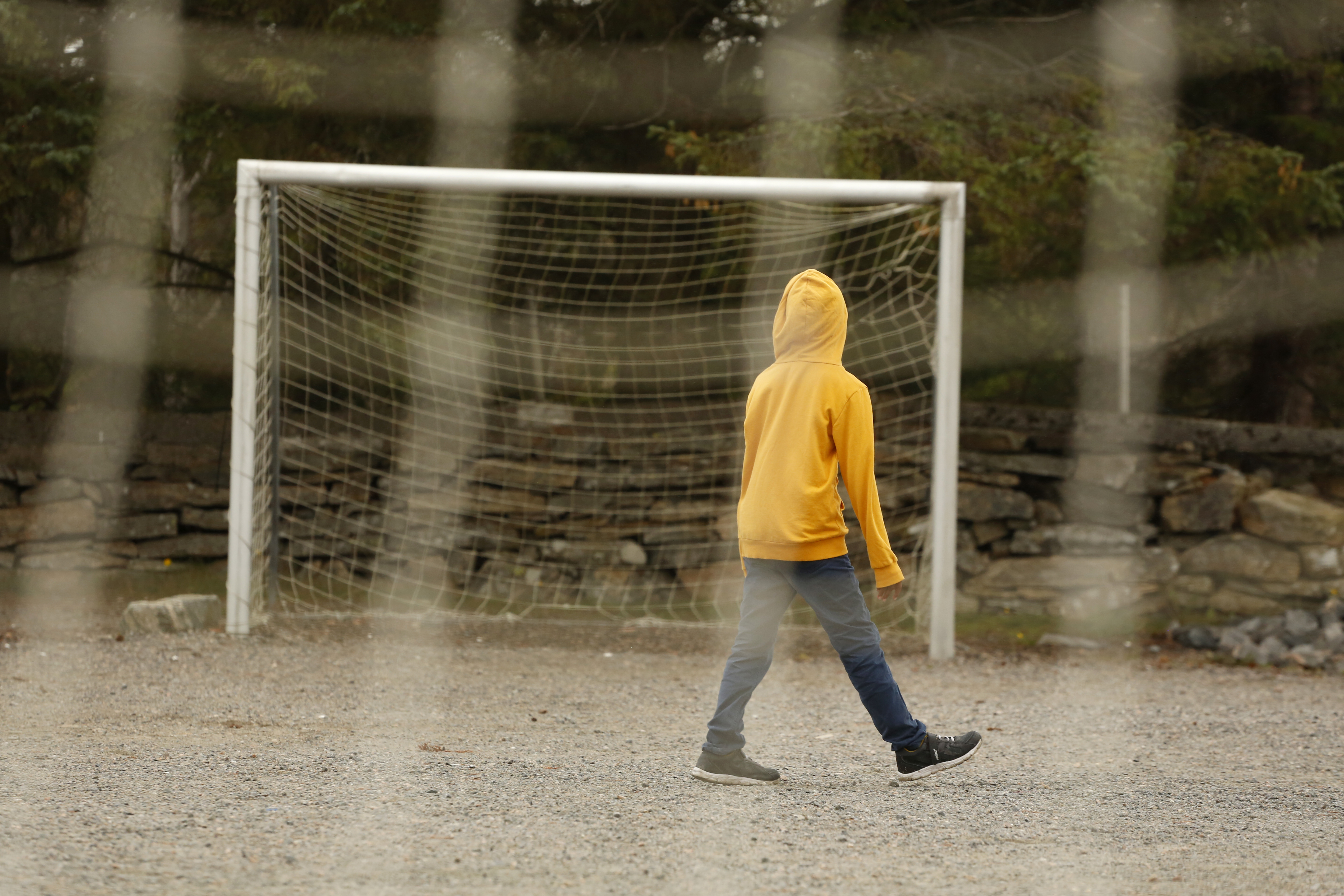 Foto: Barn i gul hettegenser går ute på en grusbane like ved et fotballmål. Hetta er trukket over hodet og hen har ryggen til fotografen.
