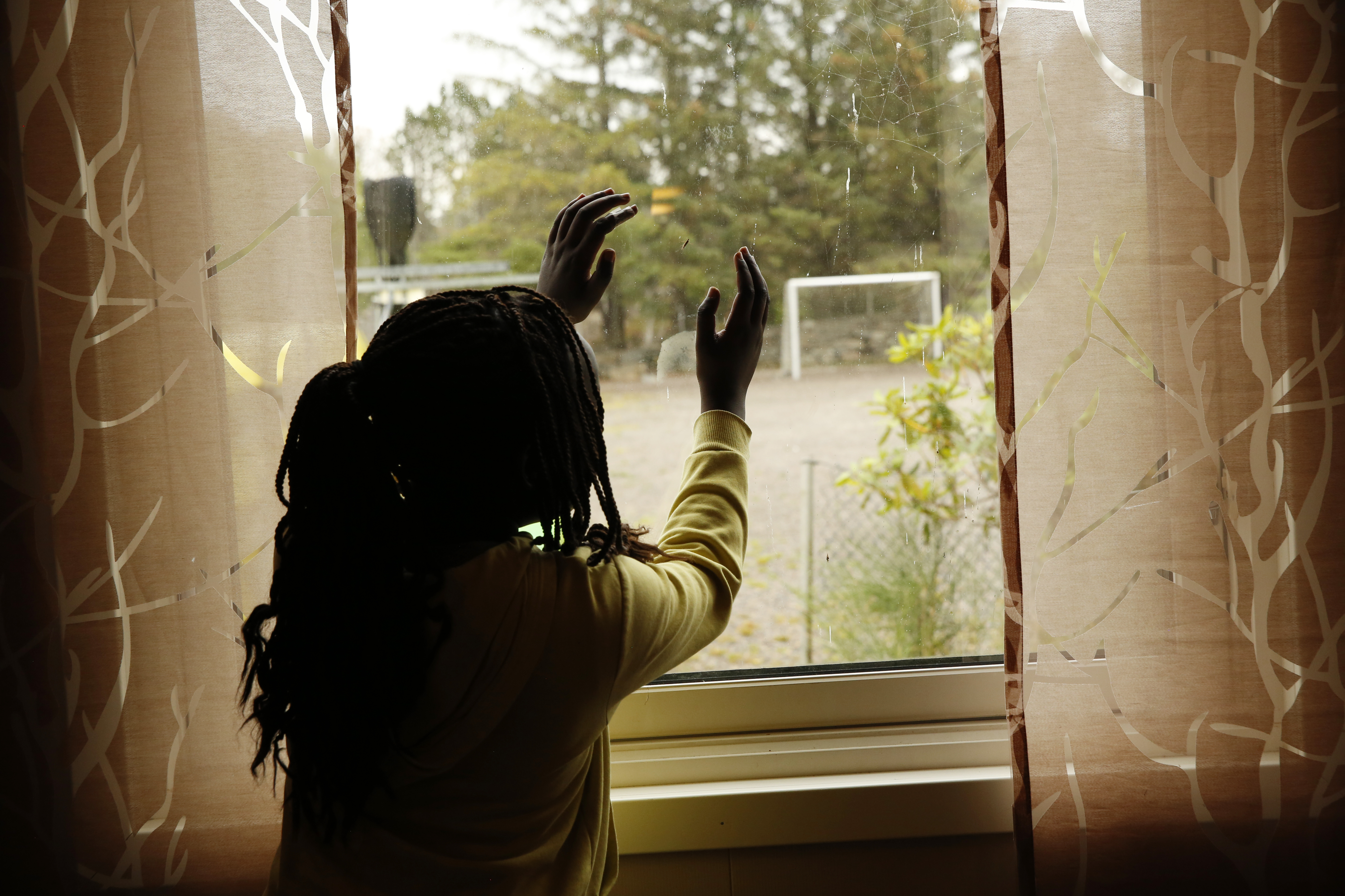 Foto: Jente med rastafletter står inne og ser ut gjennom et vindu til en grusbane med et fotballmål på. På hver side av vinduet henger det litt mørke gardiner.