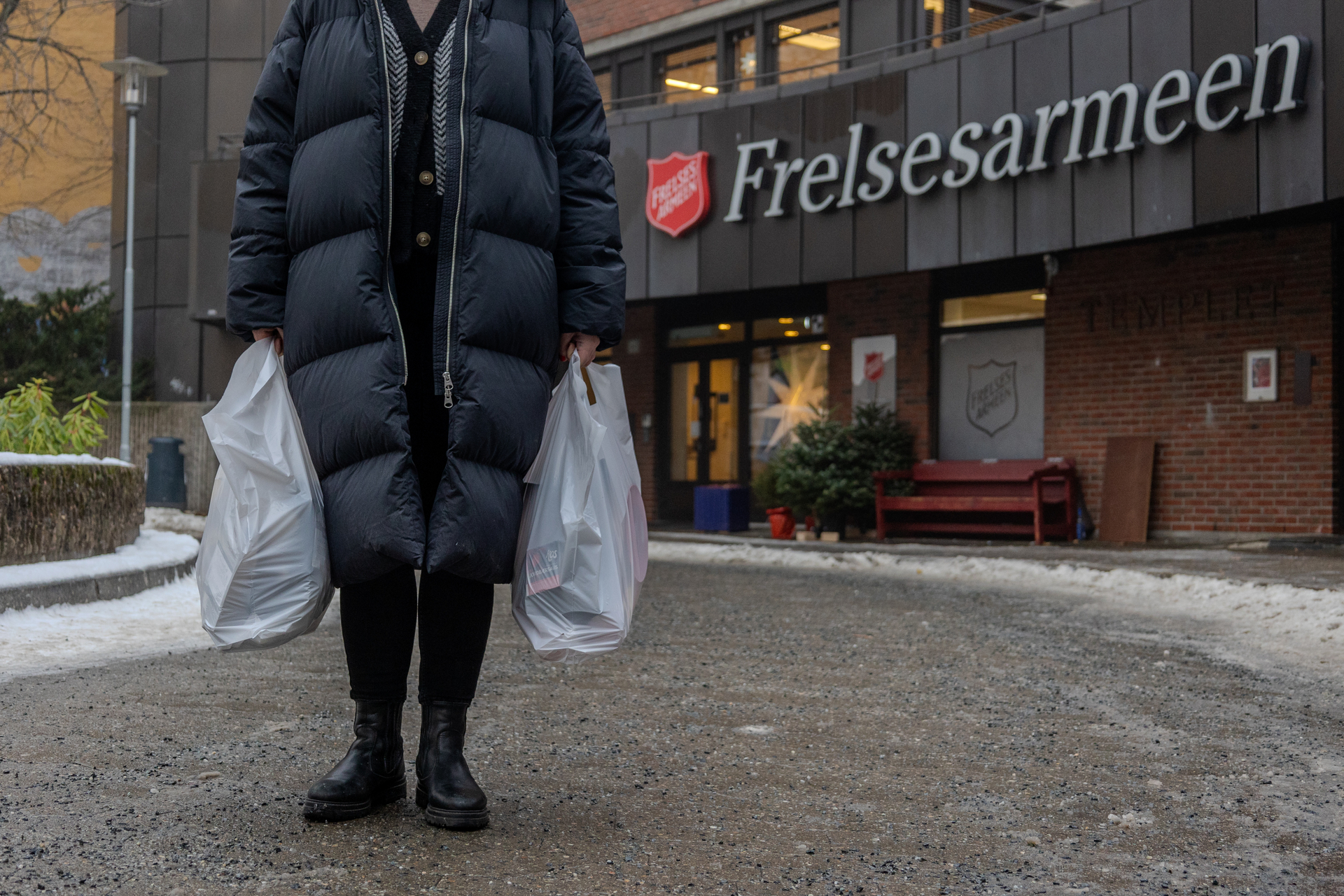 Foto: Bilde av en kvinne som bærer to poser med mat. Vi ser kun hender og føtter der hun står i en stor boblekåpe foran bygget til Frelsesarmeen.