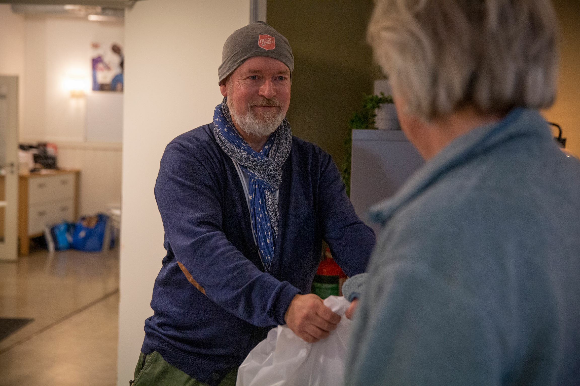 Foto: Mann med topplue med logo fra Frelsesarmeen gir en bærepose med matvarer til en kvinne.