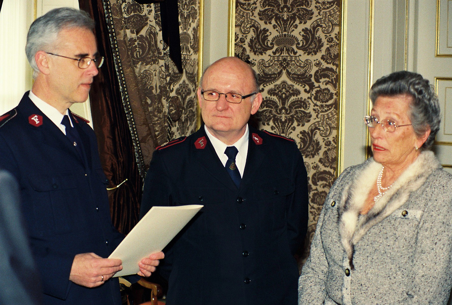 Foto: To menn med briller og i uniform fra Frelsesarmeen står ved siden av en kvinne i slutten av 60-årene med briller og oppsatt hår. Det er gulltapet på veggen bak dem. Den ene mannen holder opp et ark.