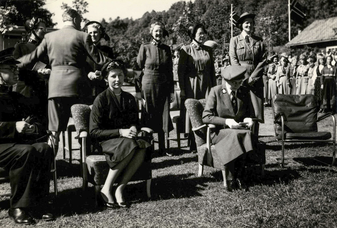 Foto: I svart-hvitt ute fra en speiderleir på 50-tallet. I en av to lenestoler sitter en ung kvinne med brunt hår. Hun har på seg en mørk drakt. Alle rundt har på seg uniformer.