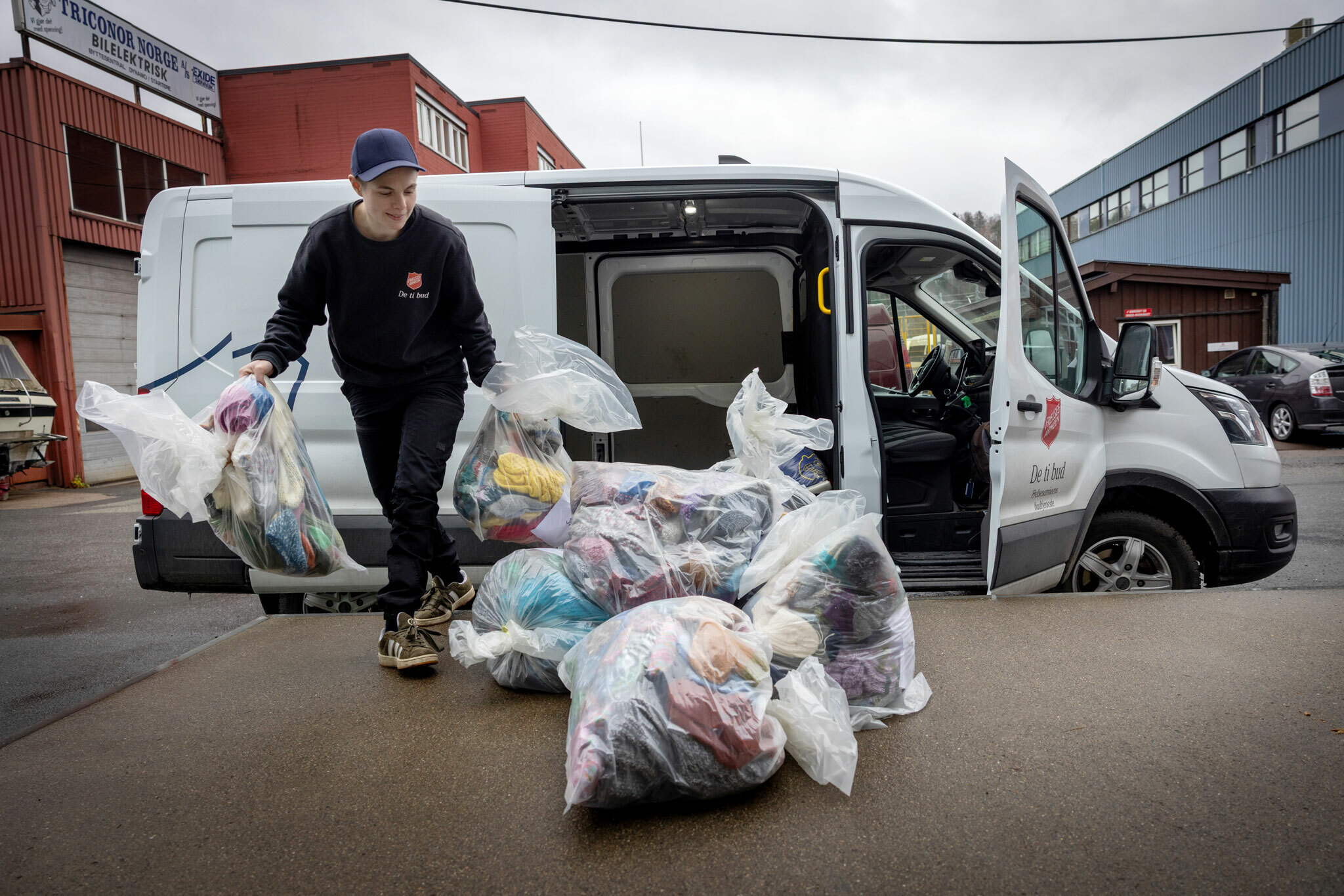 Foto: En dame med caps løfter gjennomsiktlige plastsekker med sokker. Bak henne er varebilden hun har kjørt. Den er stor, hvit og elektrisk Profilert med Frelsesarmeens skjold og navnet på budbiltjenesten: De ti bud.