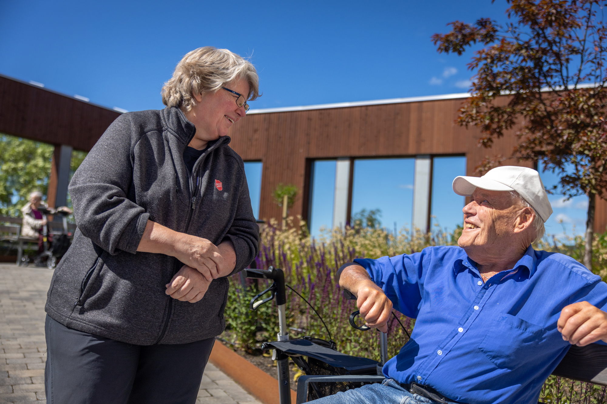 Foto: Kvinne i 50-årene med gråblondt hår og briller snakker med en eldre herre i blå skjorte og hvit caps. Han sitter på en benk i en hage. Himmelen er skyfri.