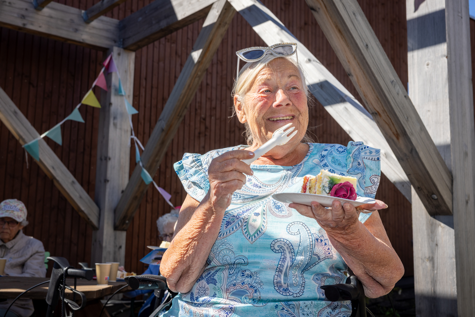 Foto: Dame i 80-årene med et par hvite solbriller på hodet spiser marsipankake og smiler i sola.