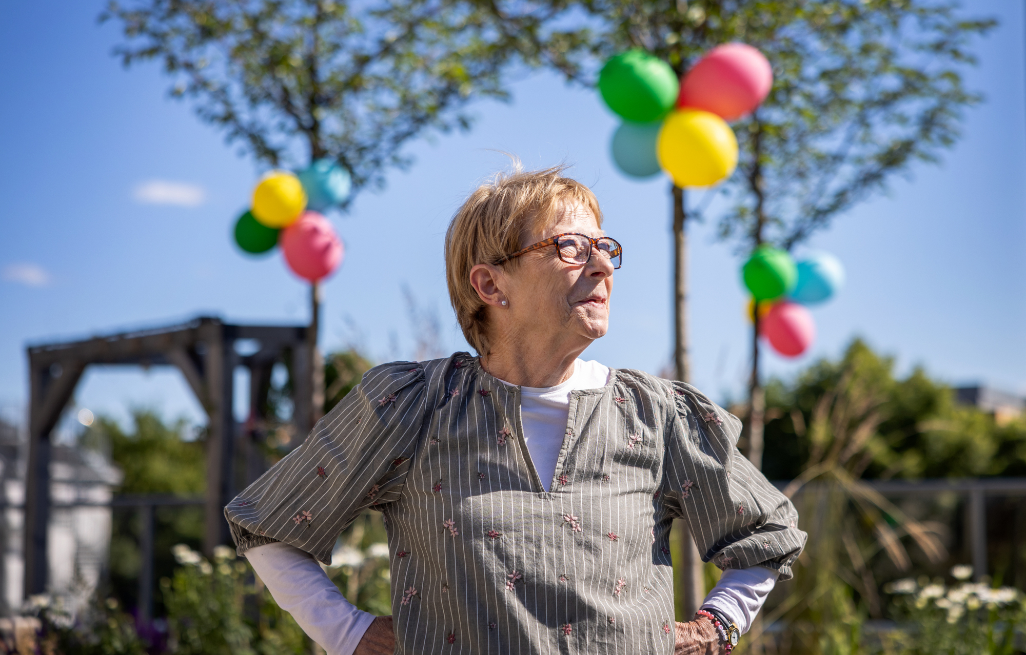 Foto: Kvinne tidlig i 70-årene med briller og kort hår skuer utover- Hun har en arm i hver side og i bakgrunnen sees nyplantede trær pyntet med ballonger og en pergola.