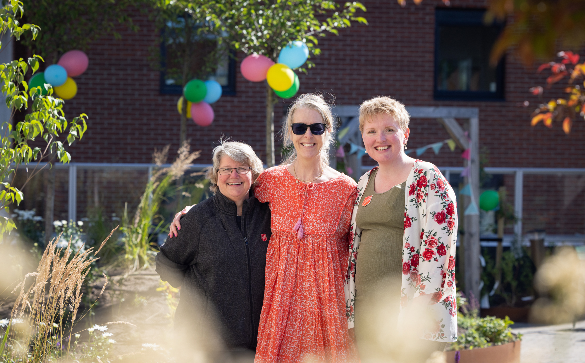 Foto: Tre damer i 50-årene står på rekke i en hage der det er festet ballonger i trærne. Det er sol og alle tre smiler og holder rundt hverandre.