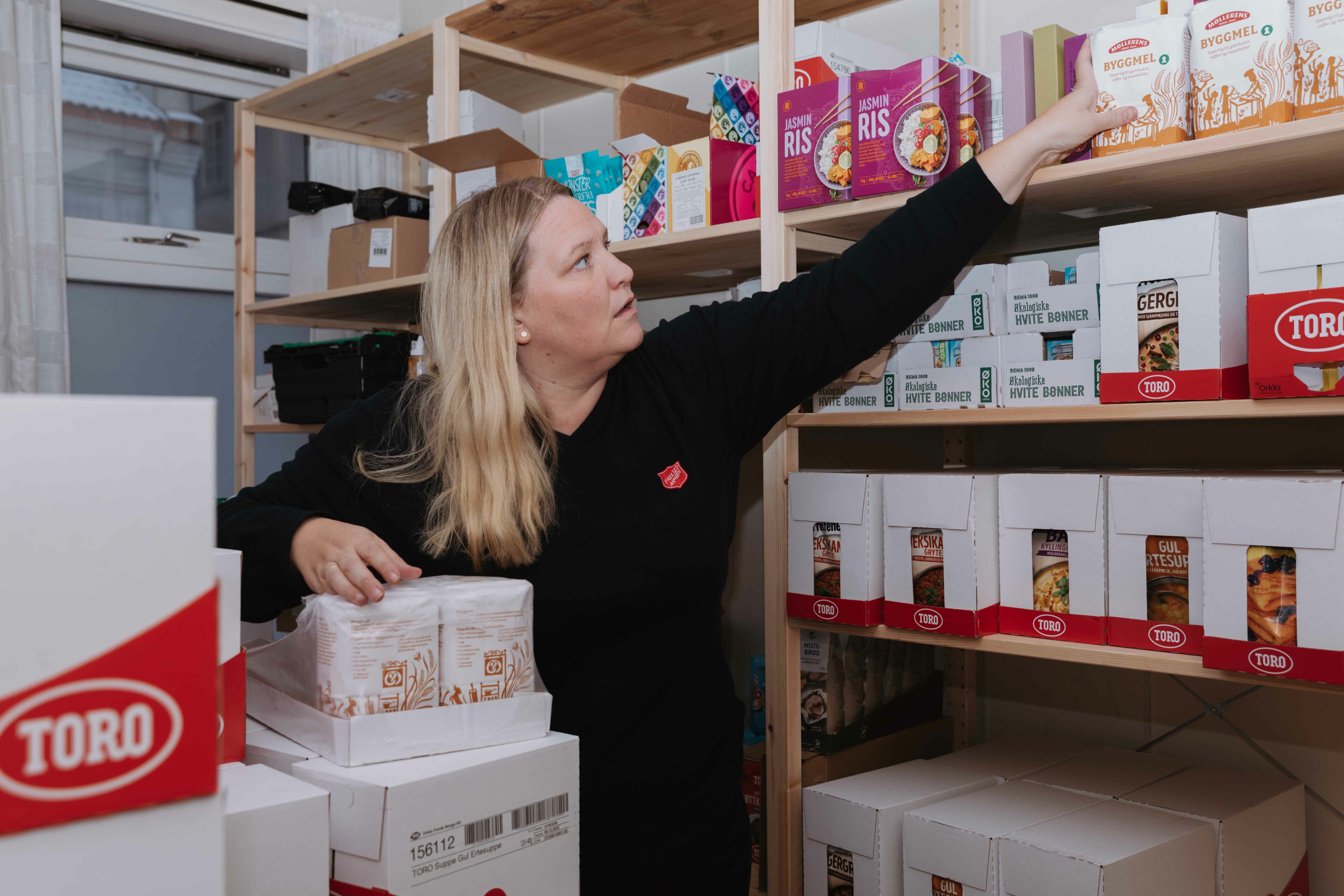 Foto: Alvorlig kvinne med langt, blondt hår strekker seg etter matvarer som står i hyller på et bakrom.