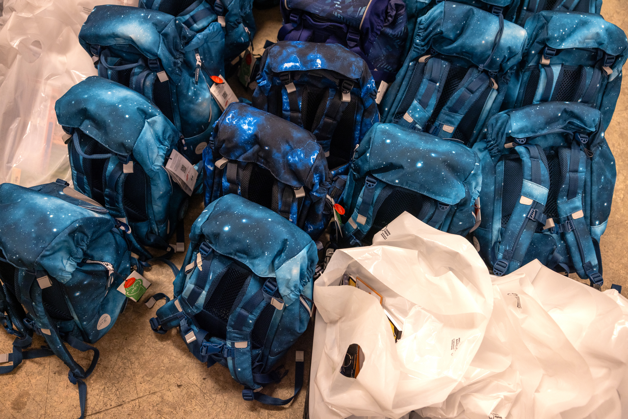 Foto: Mange skolesekker i ulike nyanser av mørkeblått står på gulvet. I. ett hjørne av bildet sees hvite plastposer fylt med ting.