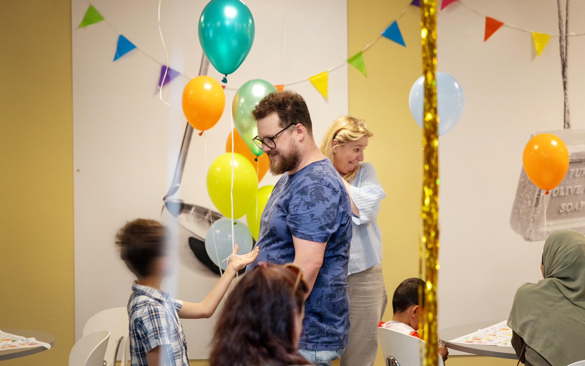Foto: En ung mann med blå t-skjorte, kort brunt hår og briller deler ut en ballong til en liten gutt som står med ryggen til. I bakgrunnen står en ung kvinne i hvit topp og snakker med en annen gutt og en mamma med brun hijab, begge med ryggen til.