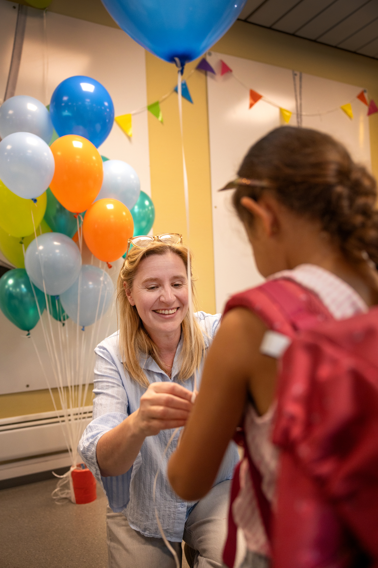 Foto: En ungkvinne med lyst, langt hår sitter på huk og smiler. Hun knyter snora til en blå ballong rundt håndleddet til ei jente, som står med ryggen til med sekk på ryggen.