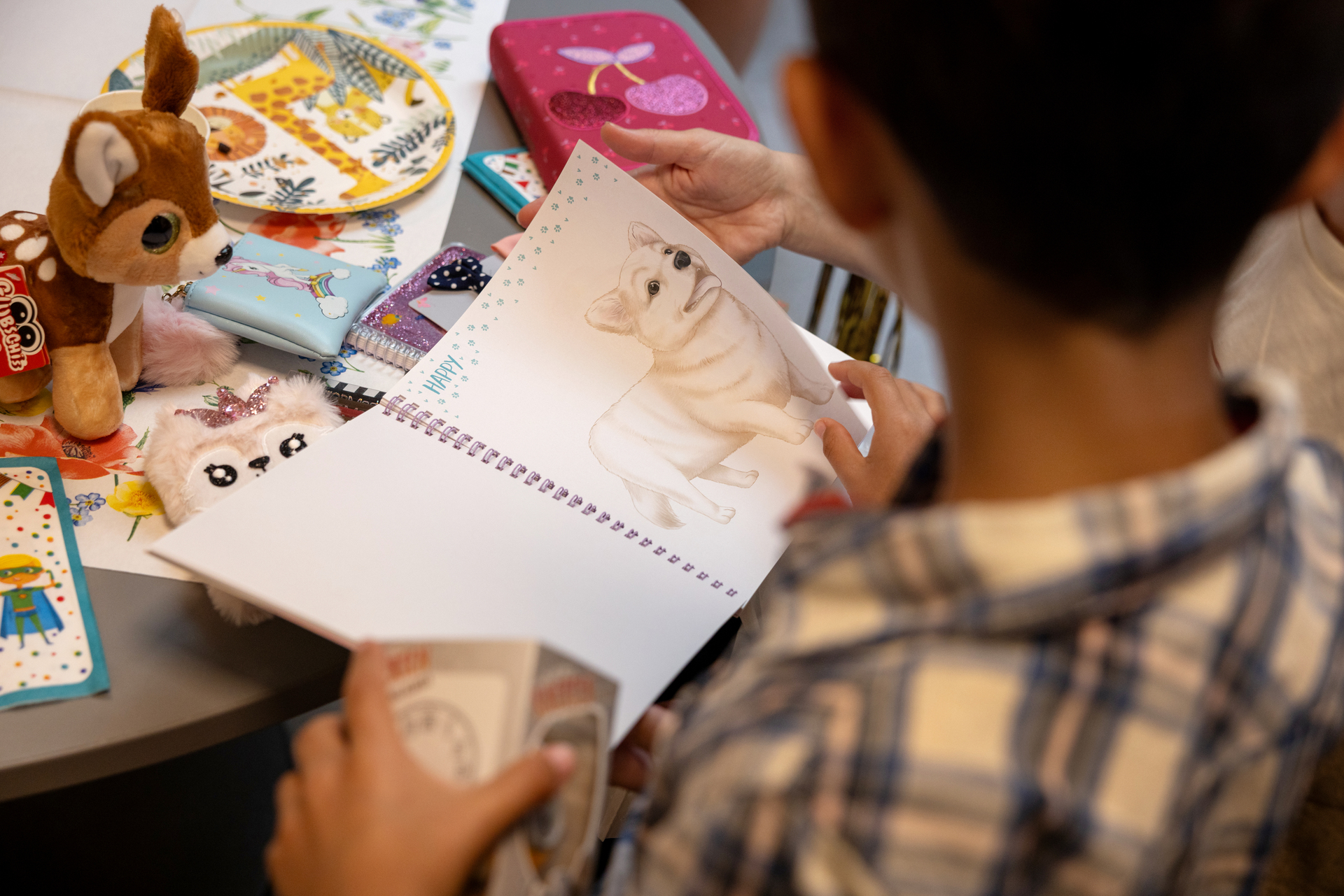 Foto: En gutt med mørkt kort hår og rutete skjorte sitter ved et bord, han sees bakfra. O den ene hånden holder han en hvit eske, med den andre blar han i en bok, han har oppe en side med en hund som kan fargelegges. På bordet foran ham står et kosedyr, pennal og mange andre småting.
