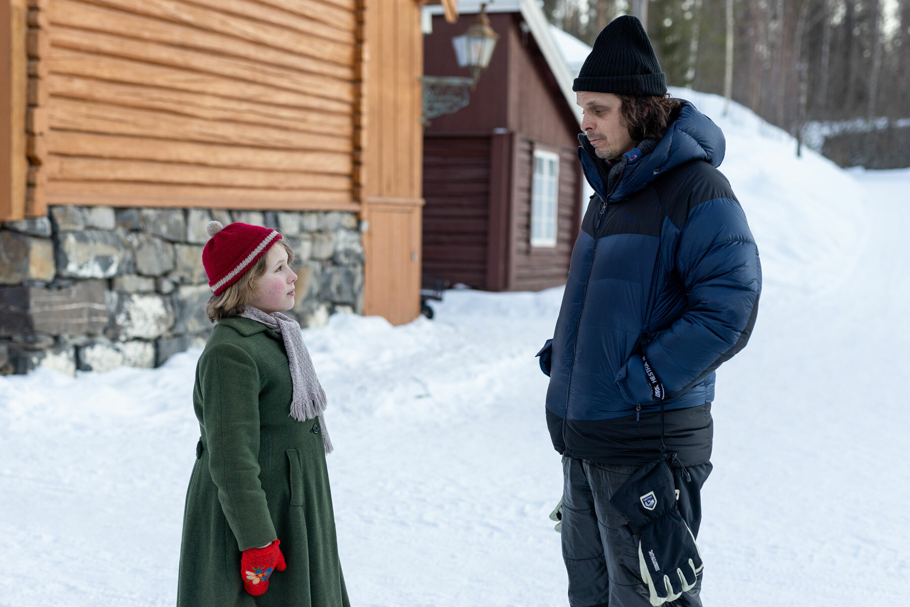Jente kledd i gammeldagse klær snakker sammen med en mann med topplue og boblejakke. Mellom husene er det snø.