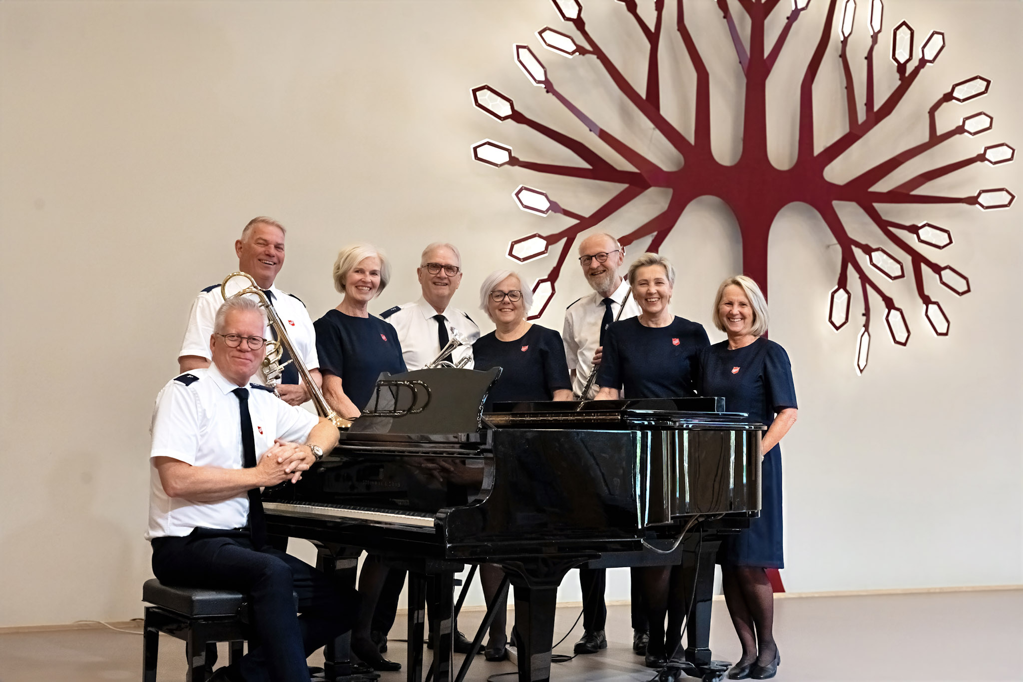 Foto: Fire voksne kvinner og fire voksne menn står rundt et piano foran et stort kunstverk formet som et rødt tre med grener og ledlys ytterst.