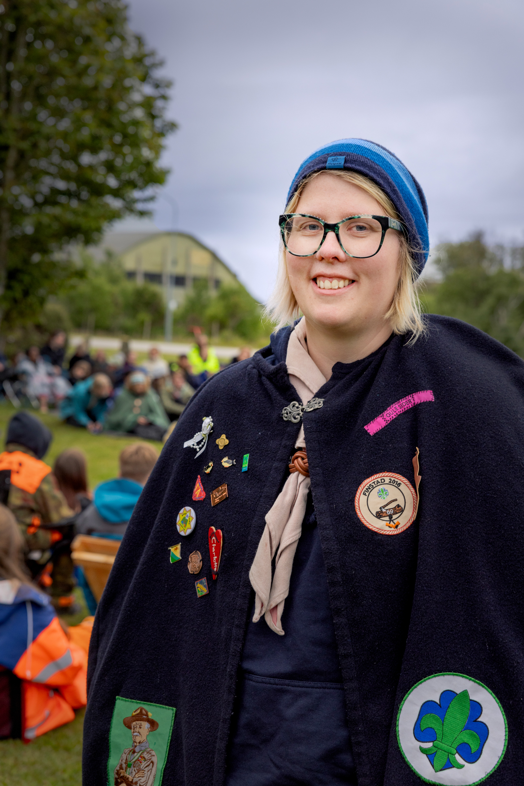 Foto: Kvinne i 30-årene med briller, speiderklær og varm kappe utenpå står foran folk som sitter på bakken.