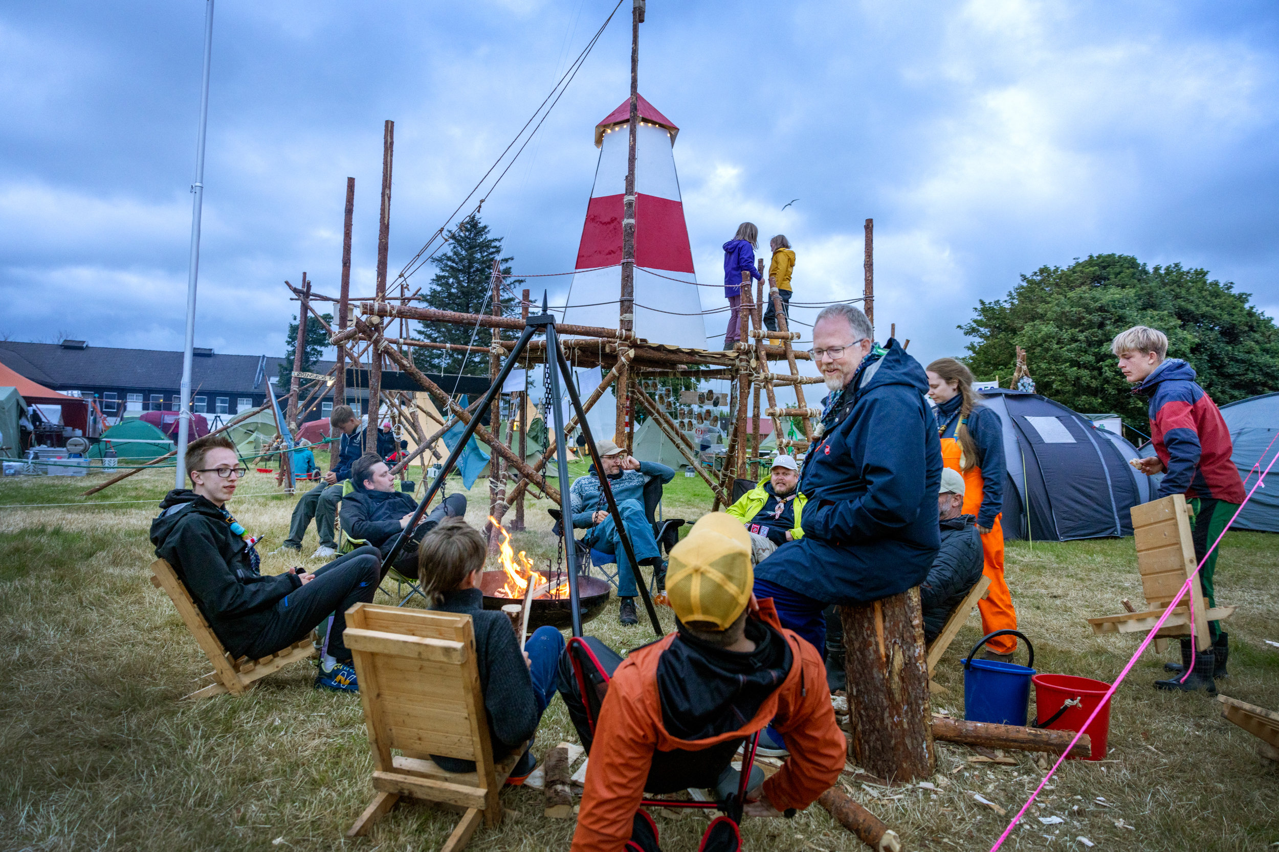 Foto: En rekke mennesker sitter litt hulter til bulter rundt bålet eller driver med andre ting like ved.