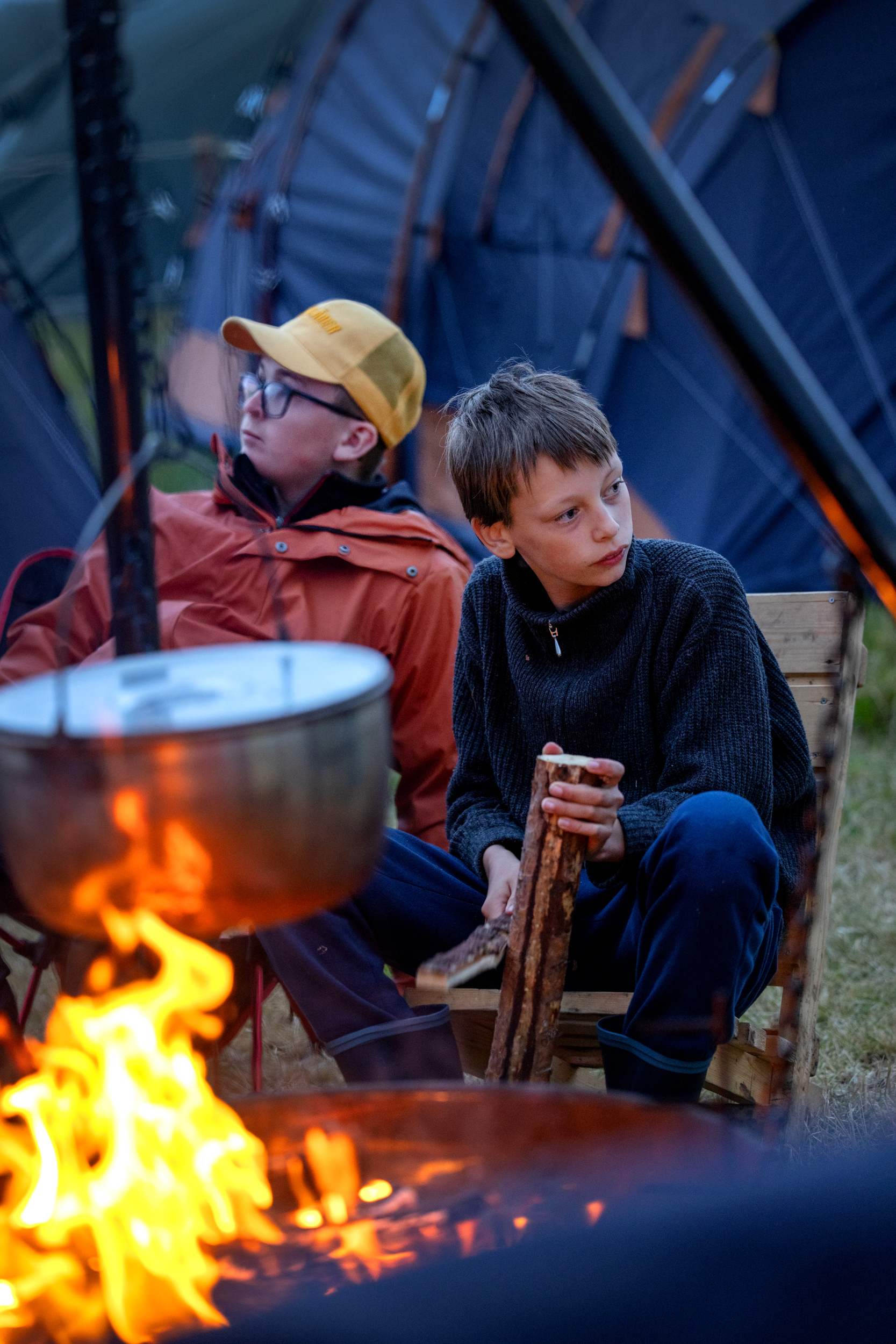 Foto: Gjennom bålpanne, der det henger en gryte ned i flammene, sitter to gutter i hver sin stol. Den ene spikker på en vedkubbe.