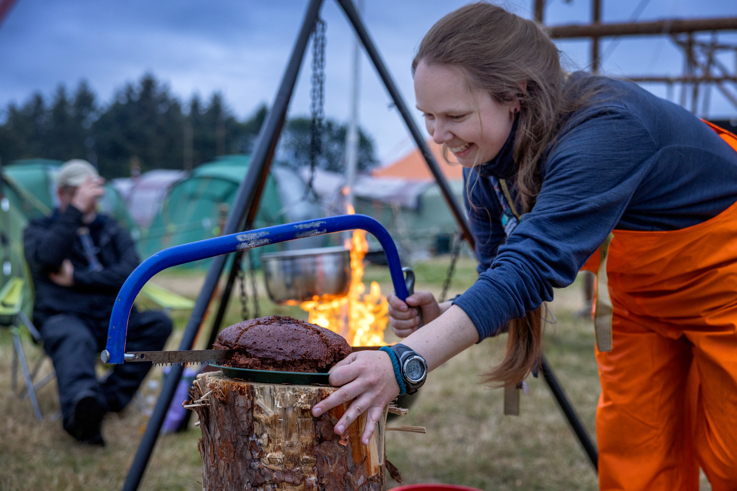 Foto: Kvinne i speiderklær og oransje oljebukse sager opp sjokoladekake med ei diger sag.