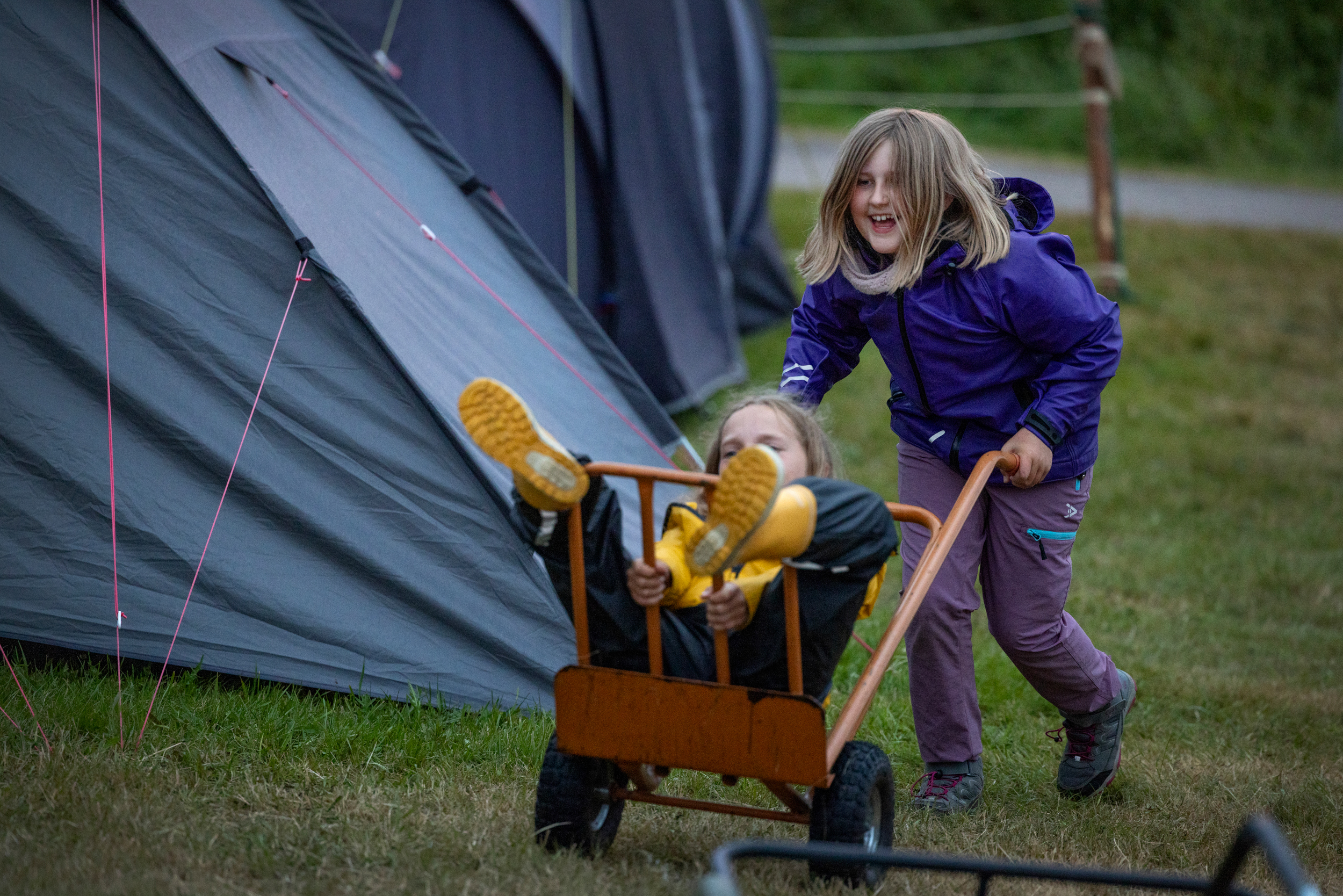 Foto: En jente i regntøy kjører en annen jente i regntøy på en sekketralle mellom teltene.