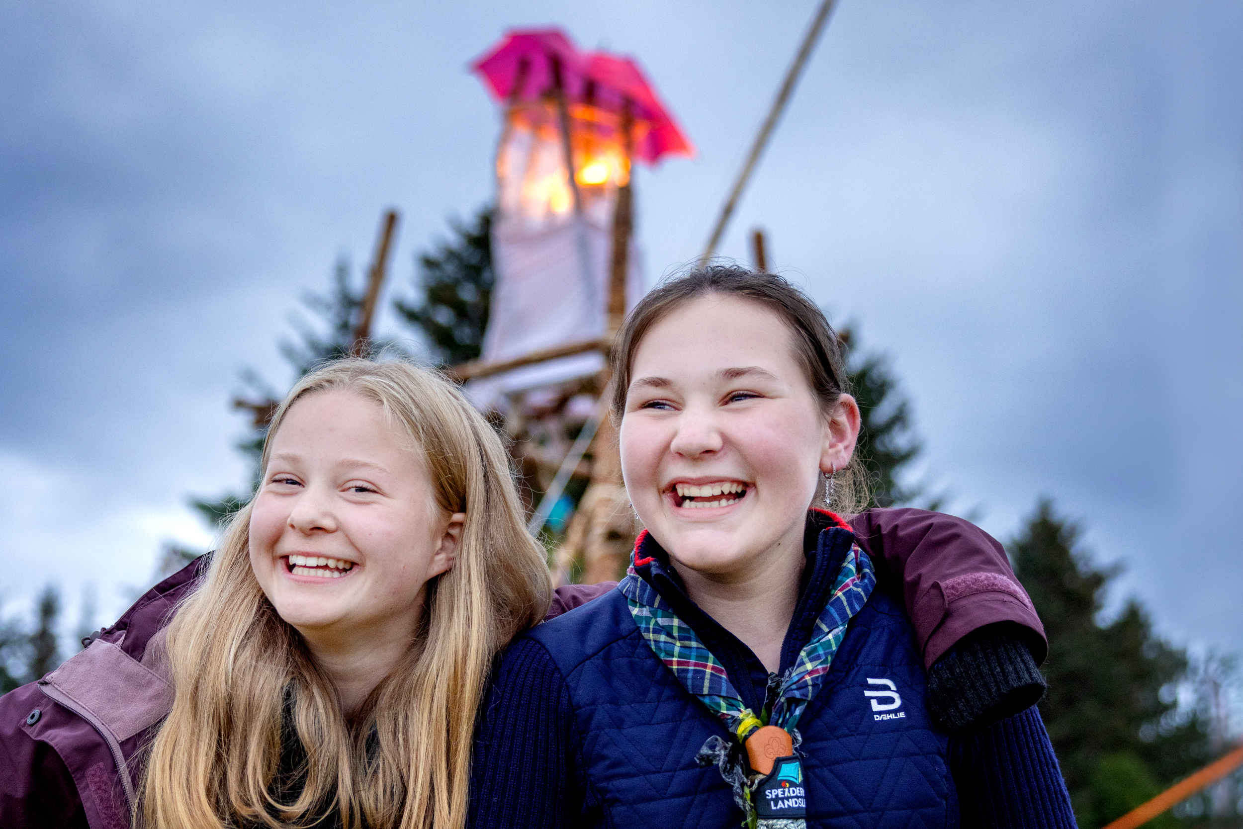 Foto: To jenter på 14 og 12 år i speiderklær smiler skikkelig. Bak dem har noen bygget et høyt tår, der toppen lyser og har en rosa paraply over som tak.
