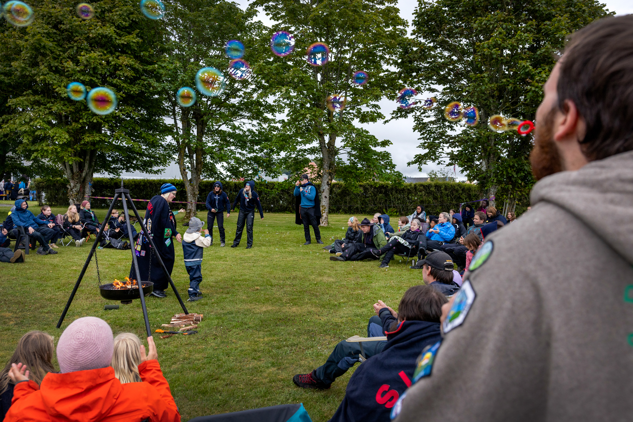 Foto: I midten en bålpanne med fyr i veden. Rundt sitter mange folk. Rett bak bålpannen går kvinne og liten sønn i regntøy.
