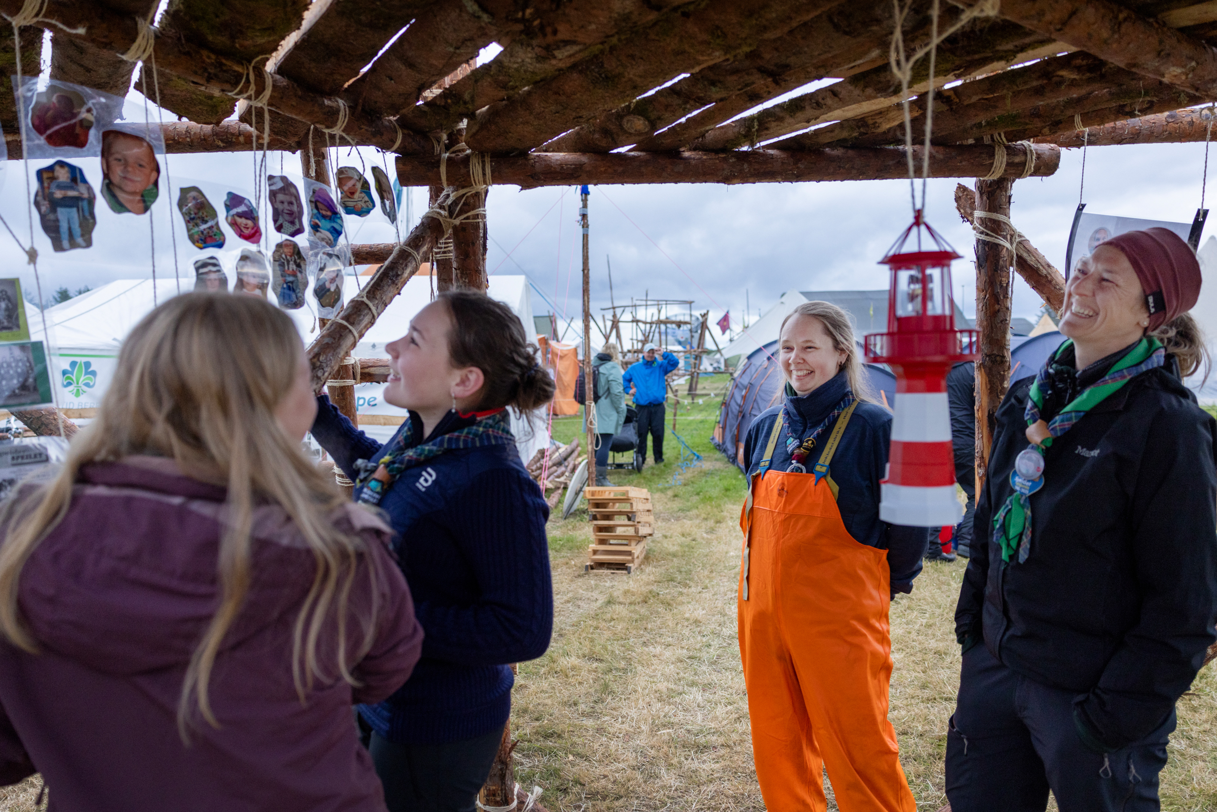 Foto: To jenter og to kvinner står inne under en portal bygget i tre på speiderleir. Her henger en "fjøslykt" formet som et fyrtårn og bilder av alle de som bor i leiren.