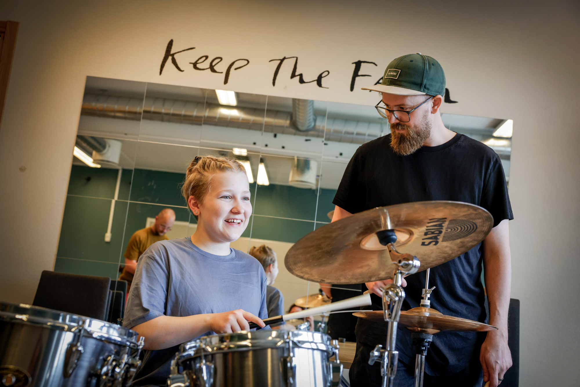 Foto: Mann med briller og caps står ved siden av en ung tenåringsjente med lyse fletter som spiller trommer.