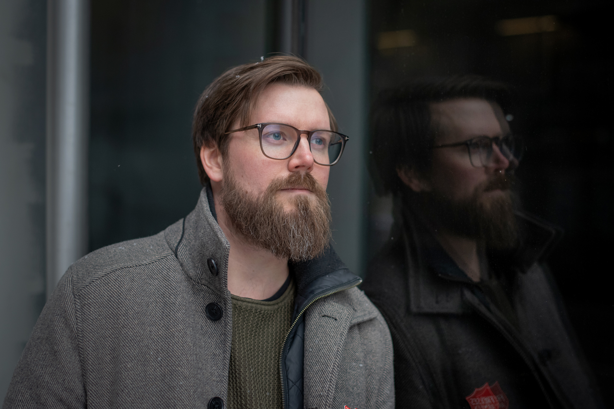 Foto: Mann i 30-årene med briller, mørkt hår og skjegg ser bortover. I vinduet bak ser man refleksjonen hans. Han har på seg en beige vinterjakke.