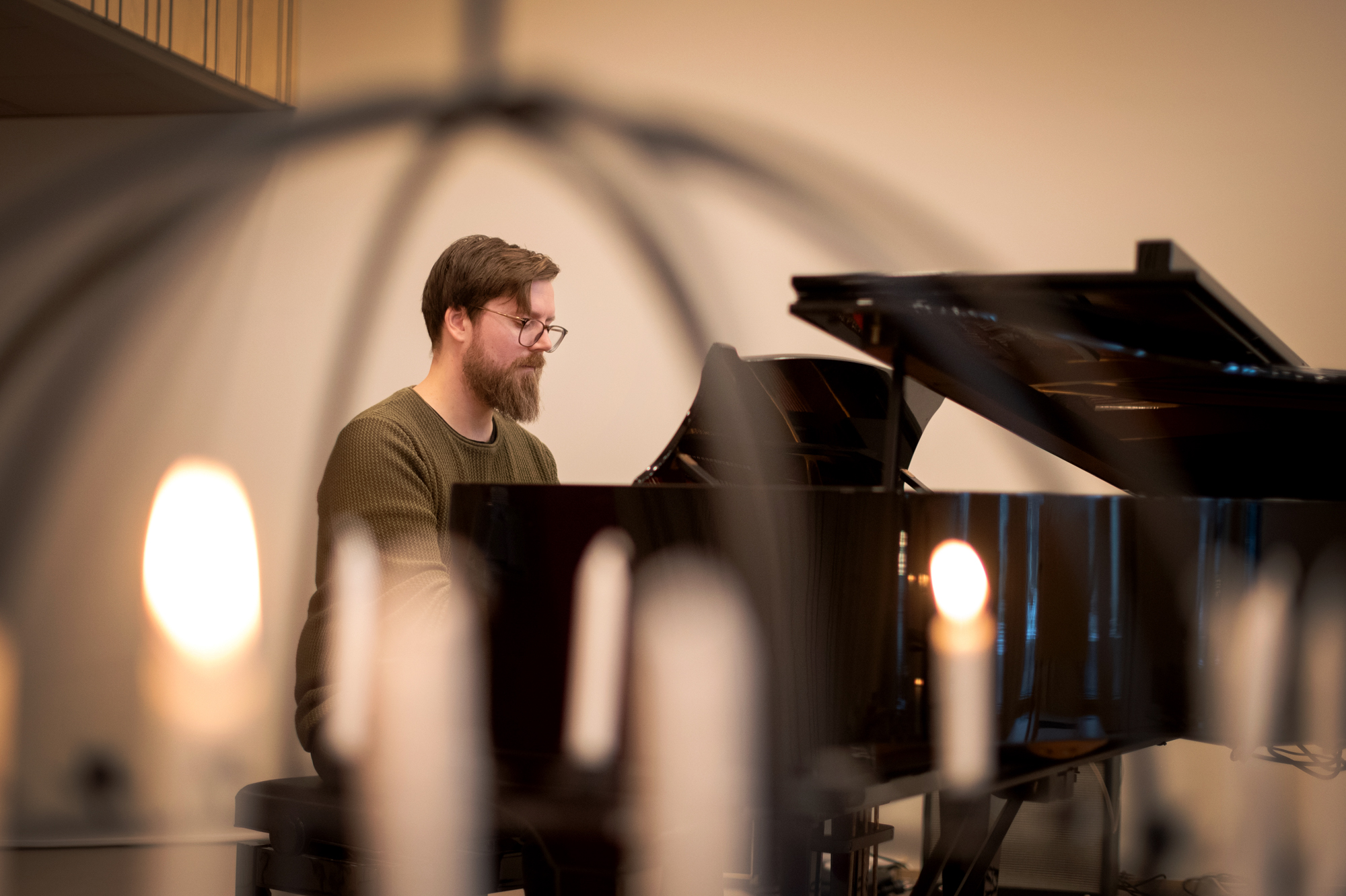 Foto: Mann med briller sitter og spiller på et flygel. Bildet er tatt gjennom en smijernsglobe med tente lys.