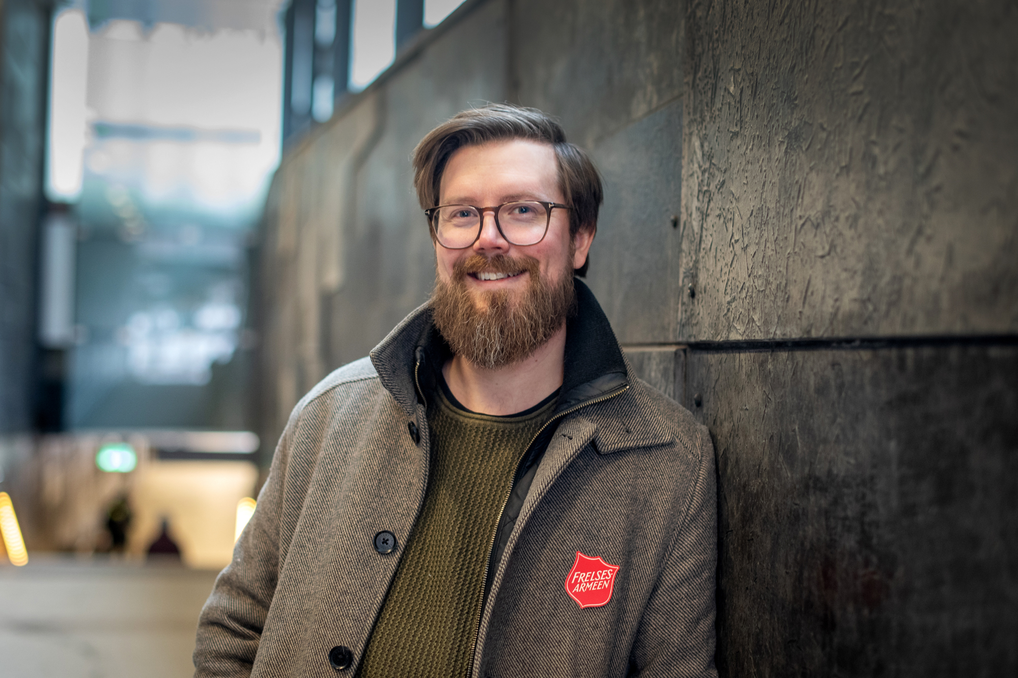 Mann i 30-årene med briller, mærkt hår og skjegg smiler mot fotografen. Han står i en jakke inntil en vegg dekket av steinfliser. På jakken har han Frelsesarmeens røde skjold.