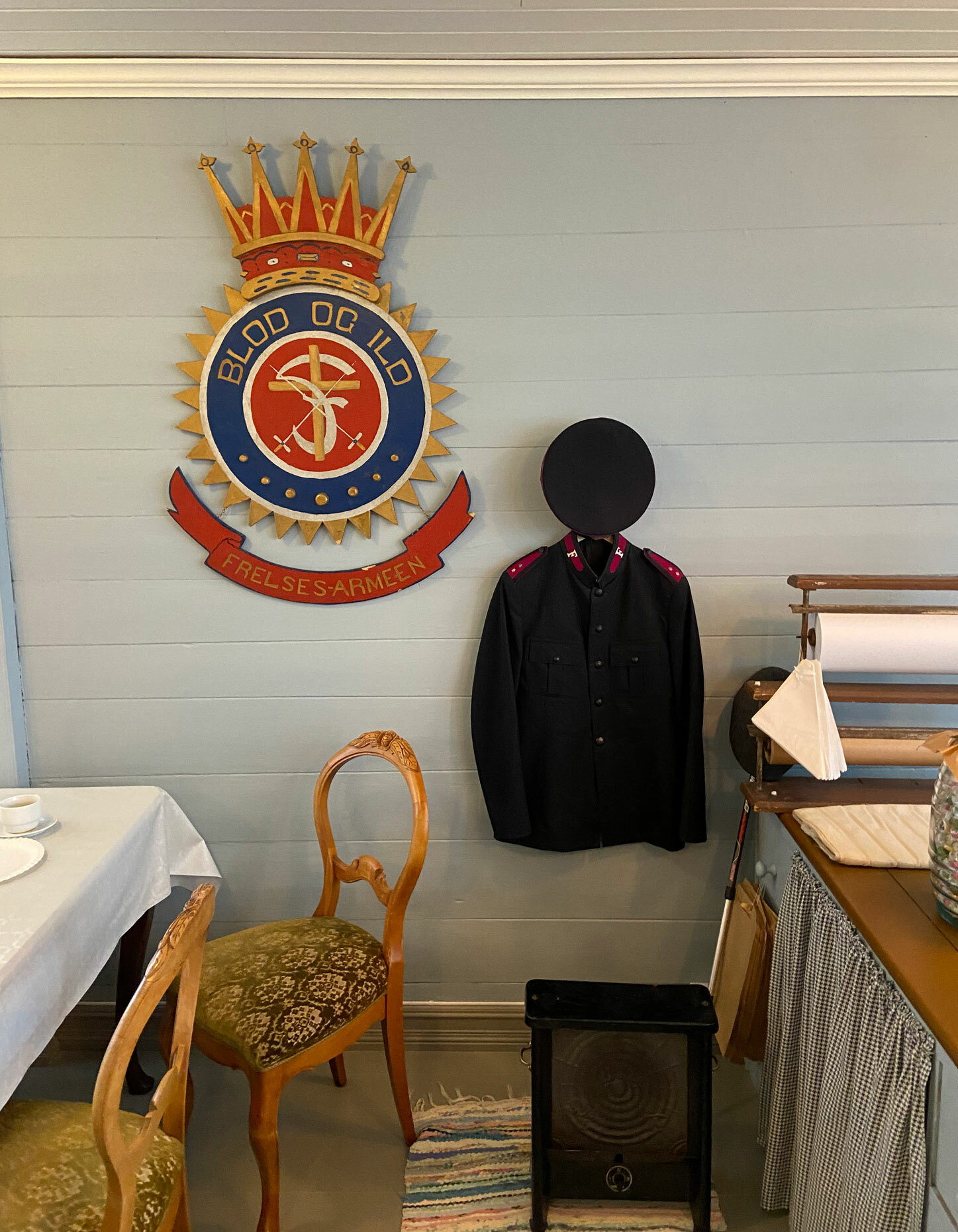 Bilde av vegg der det henger en gammel krone fra Frelsesarmeen sammen med en uniform. Gamle møbler rundt.