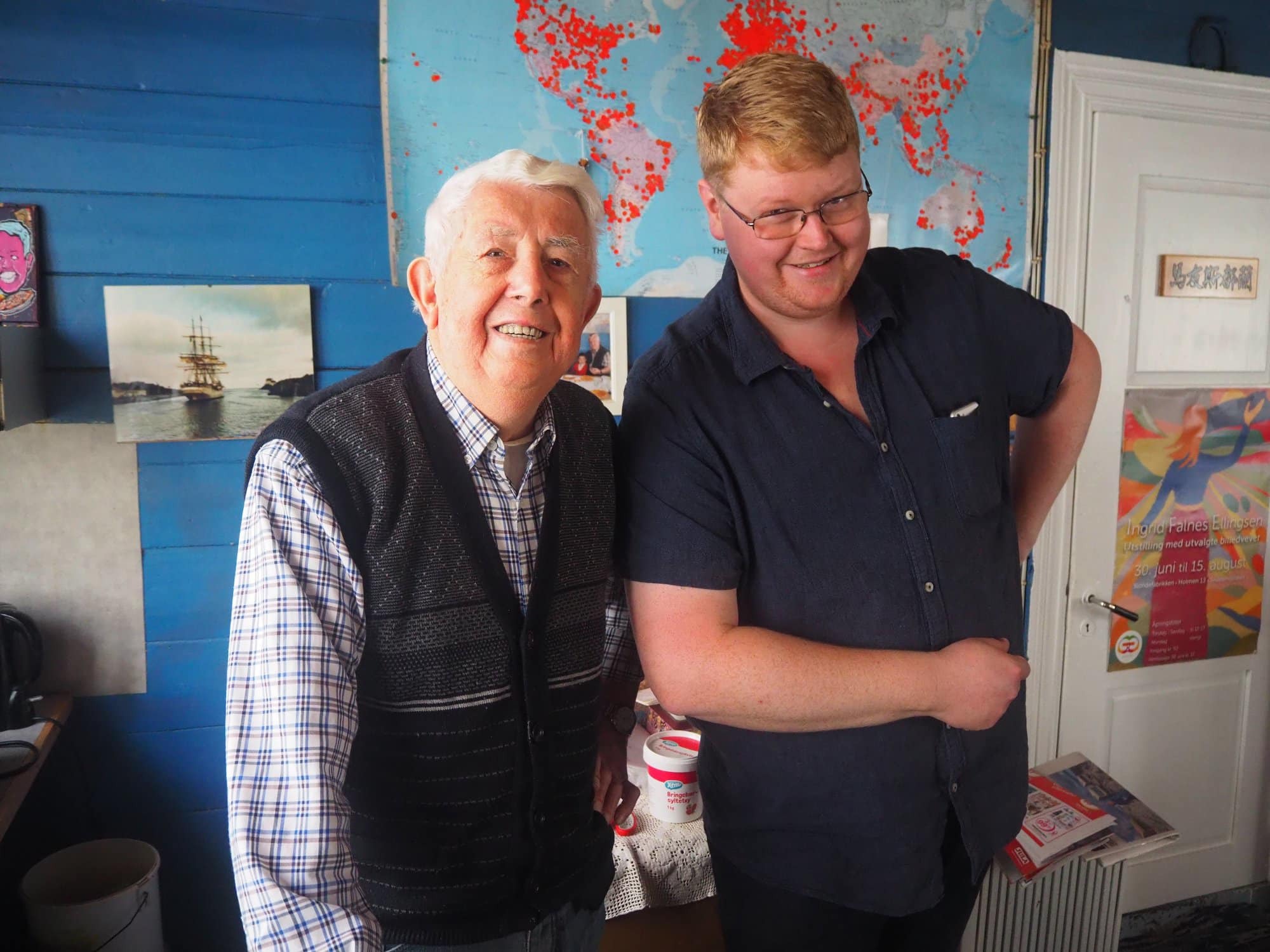 Gammel mann og ung mann ved siden av hverandre inne i et knallblått rom. På veggen bak henger et fargerikt verdenskart.