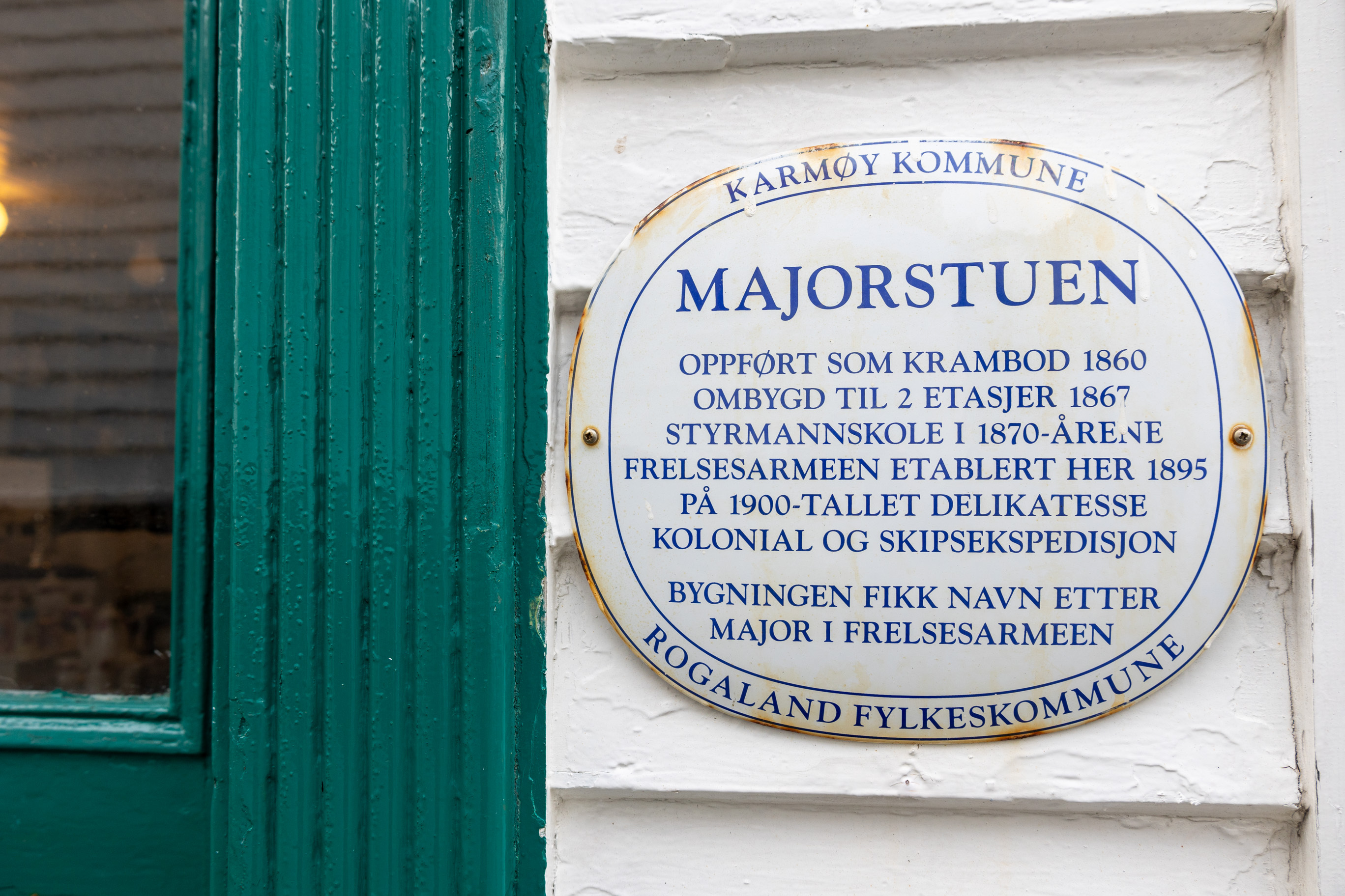 Hvitt, gammelt porselensskilt med blå skrift. Henger på ytterveggen ved inngangsdøra.