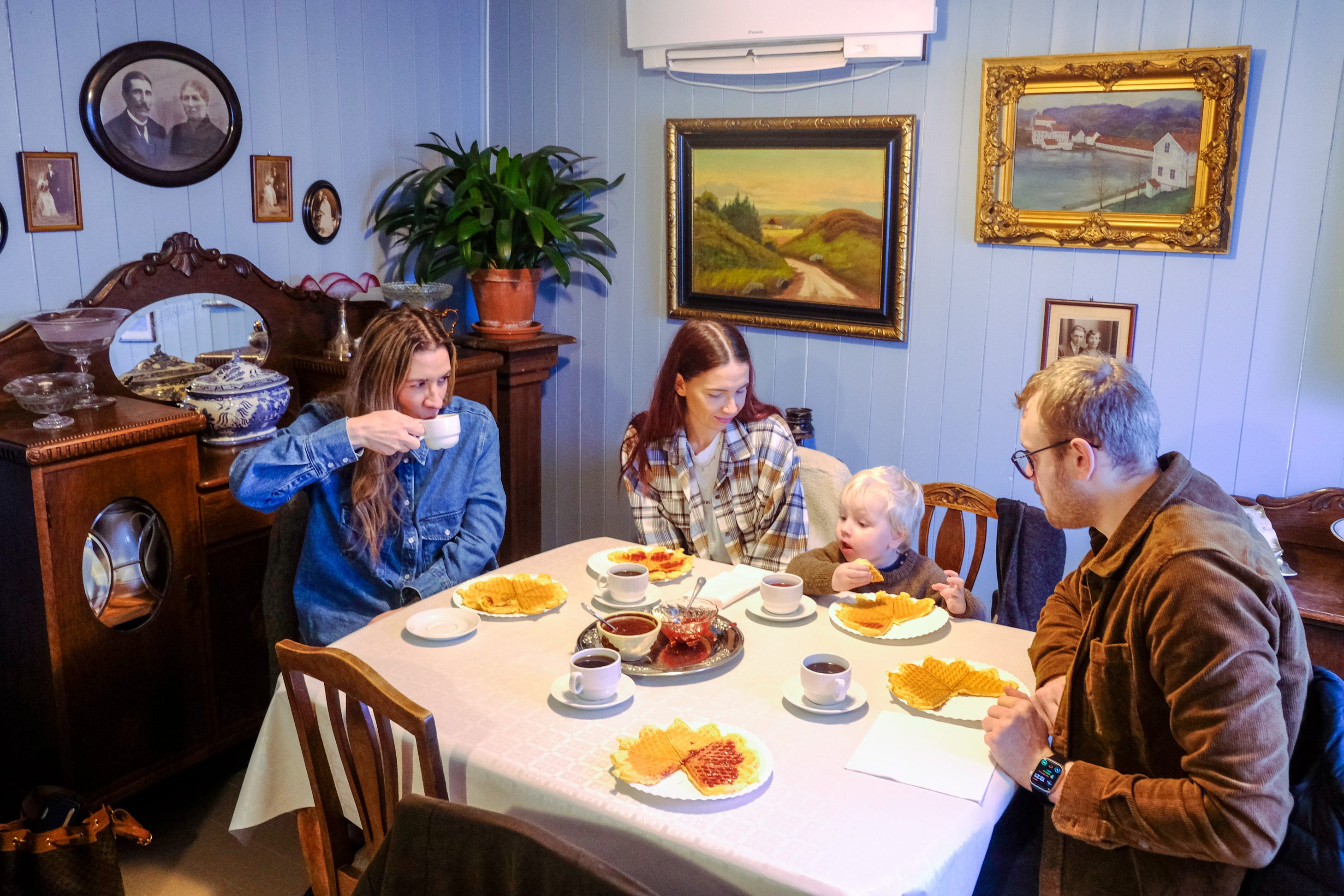 Tre voksne og et lite barn rundt et bord spiser alle vafler i det som er en gammel spisestue med tilhørende skjenk.