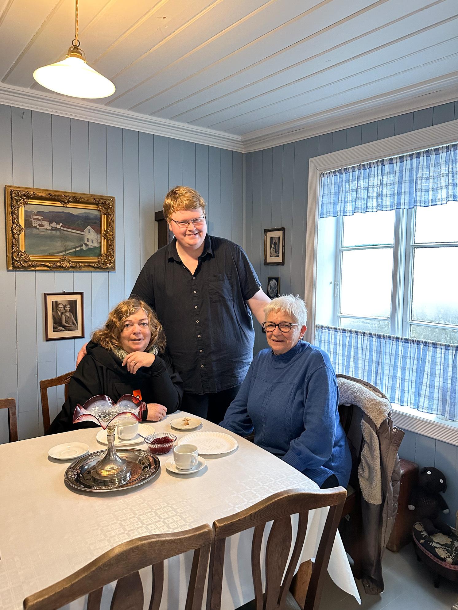 Yngre mann står mellom to voksne damer som sitter ved et gammelt bord med duk på.